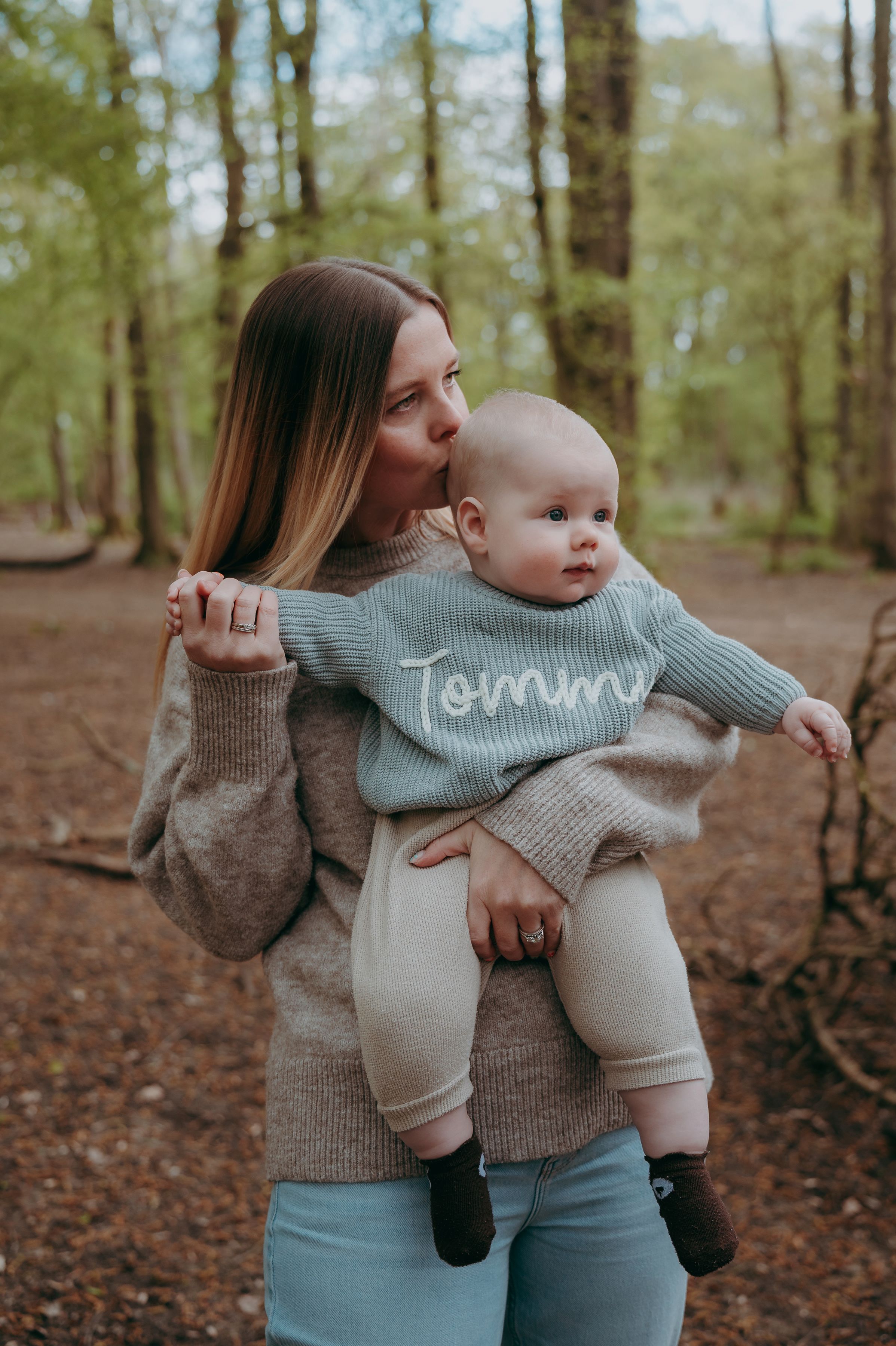 Mother kissing baby named Tommy in forest, baby wearing sage green knit sweater with name embroidered.