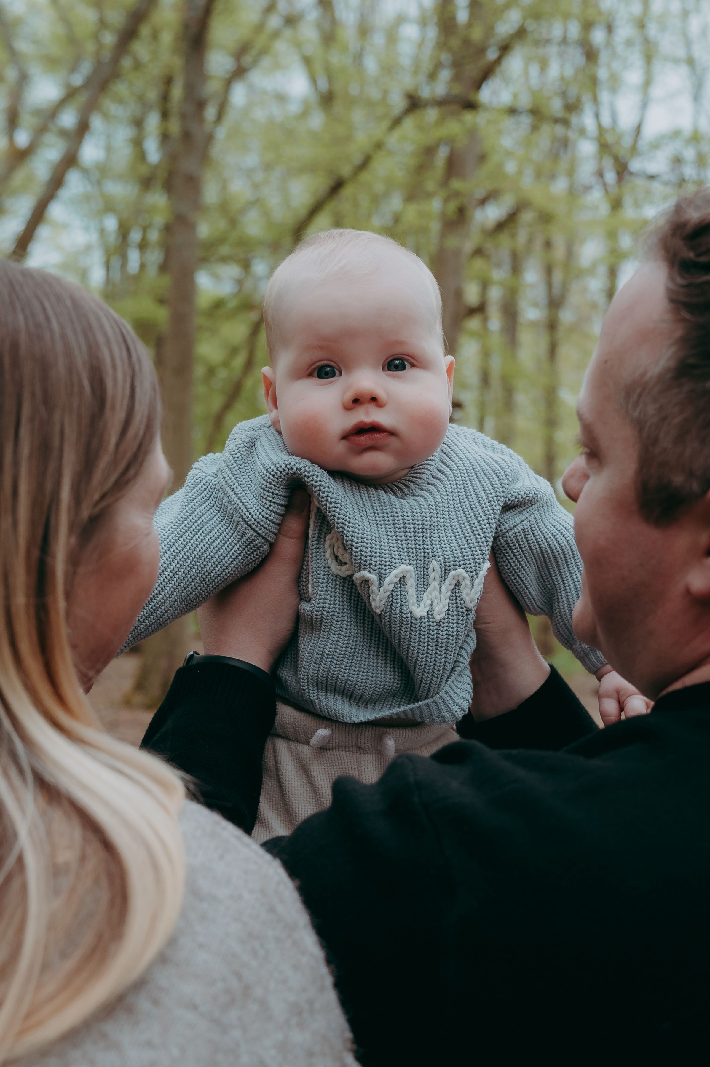 Parents lifting their baby in a green sweater outdoors in a forest setting.