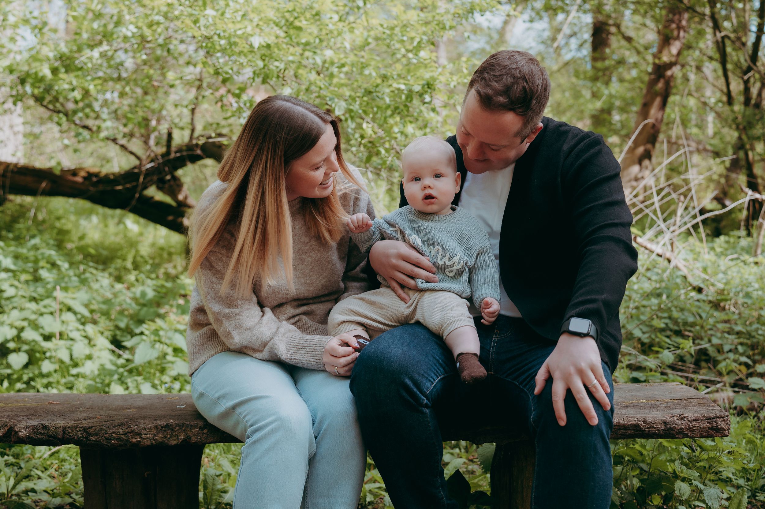 A happy young family with a baby sitting on a wooden bench surrounded by lush green trees outdoors.
