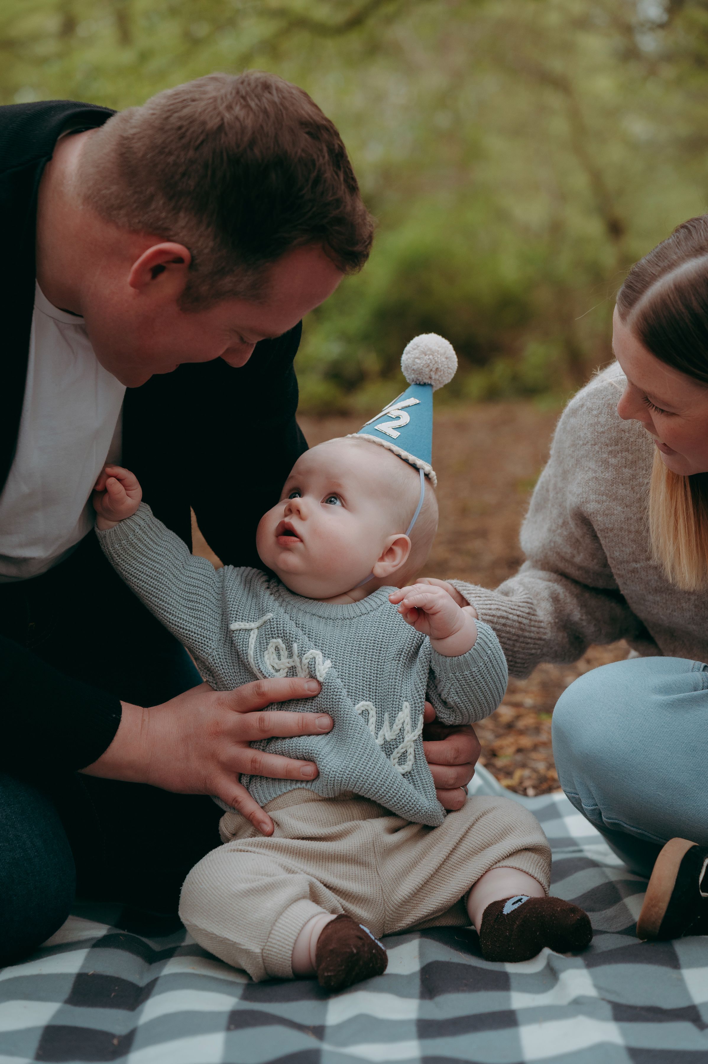 A baby in a blue party hat and knit sweater sits on a blanket outdoors, celebrated by two smiling parents.