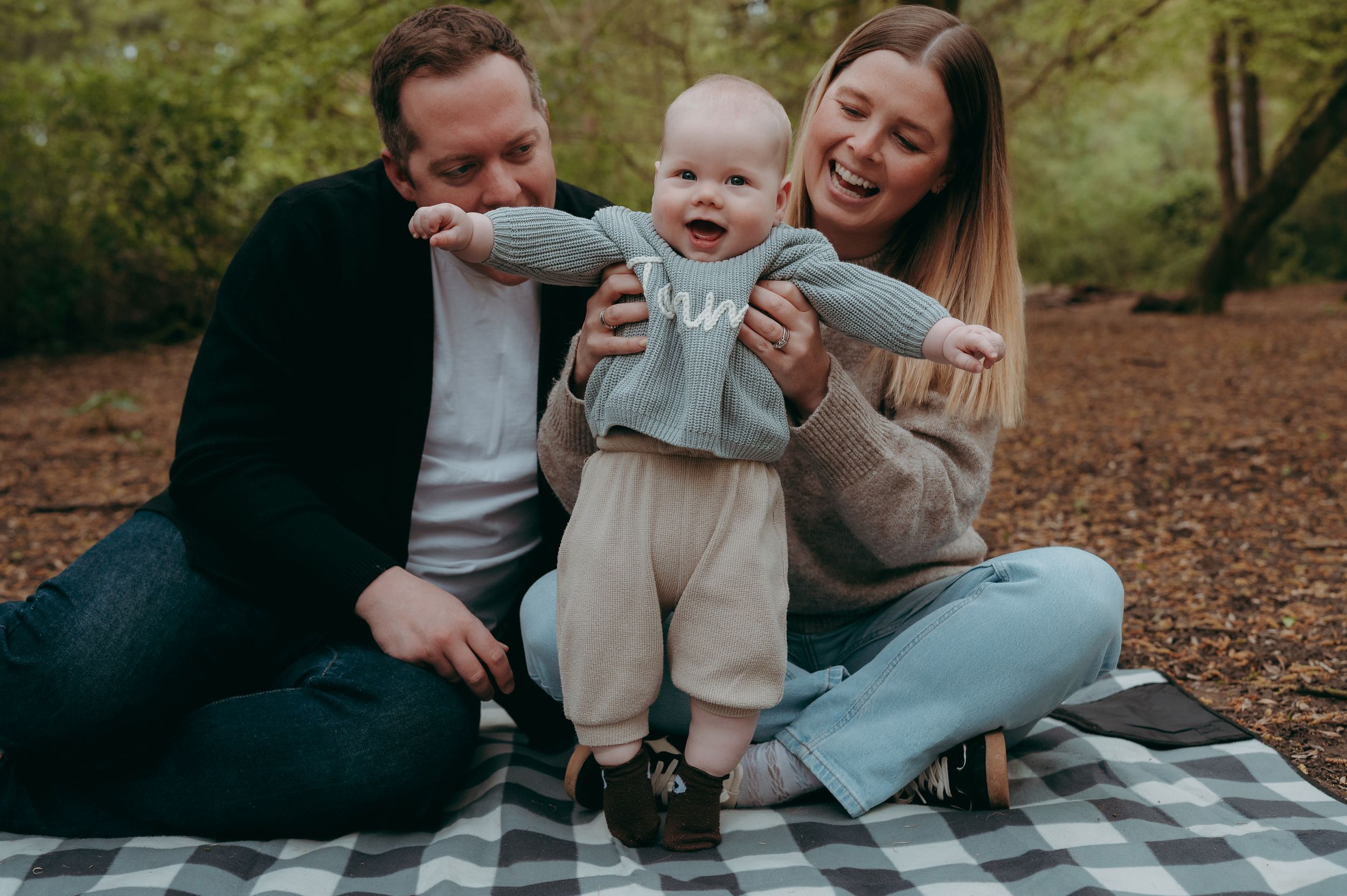 Happy family with laughing baby sitting on plaid blanket in autumn woodland setting.