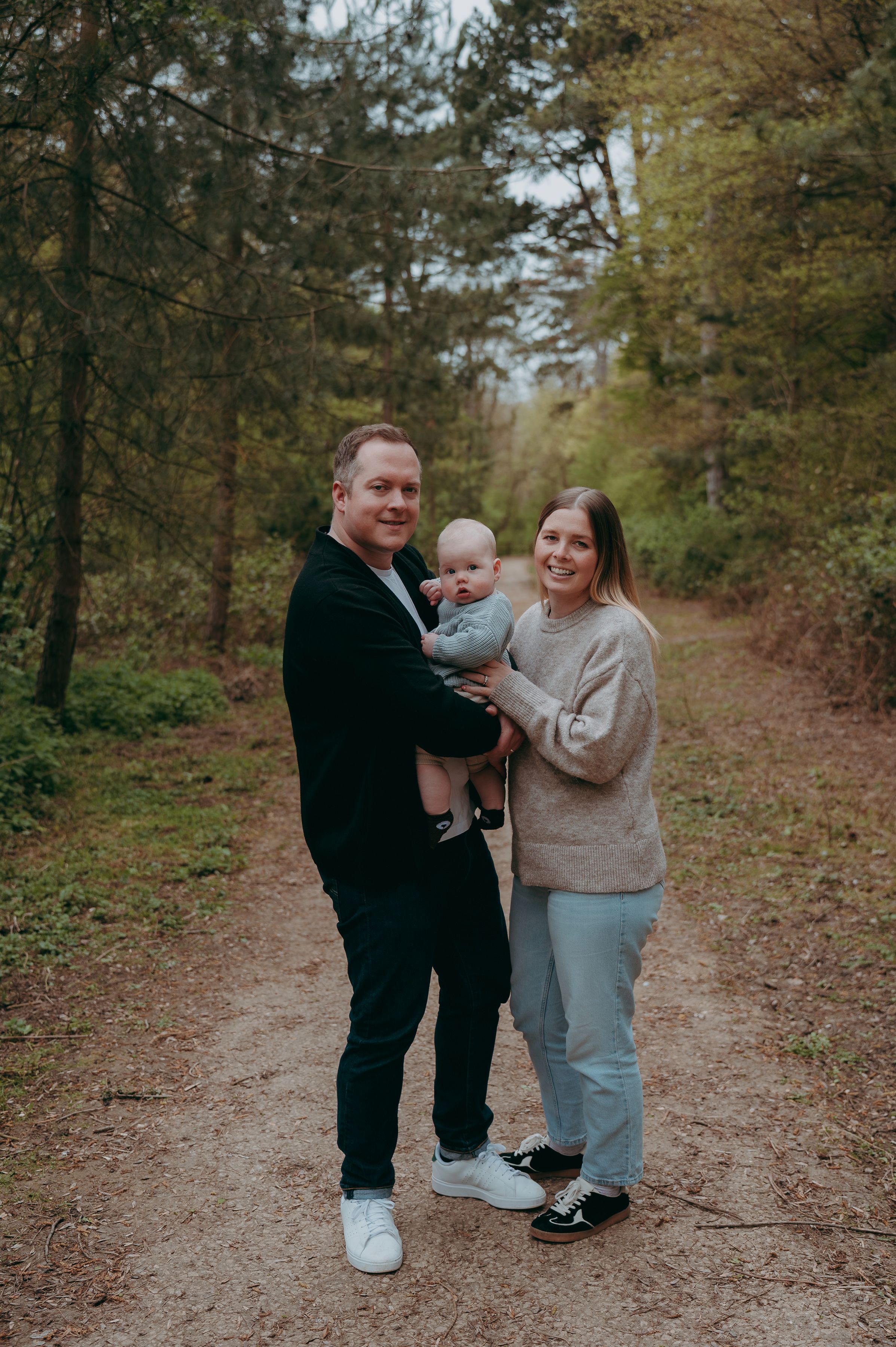 Happy family with baby posing on a woodland path surrounded by trees.