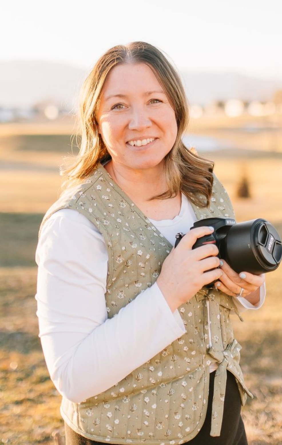 A photographer in a floral vest and white sweater holds a professional camera outdoors during golden hour.