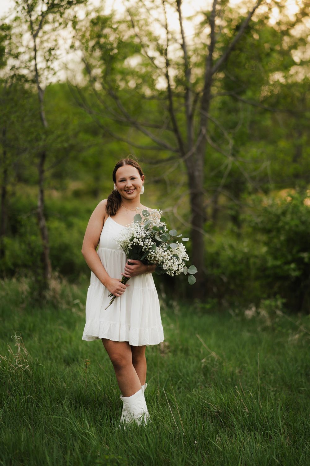 A smiling young woman in a white dress and boots holding a white wildflower bouquet in a lush green field.