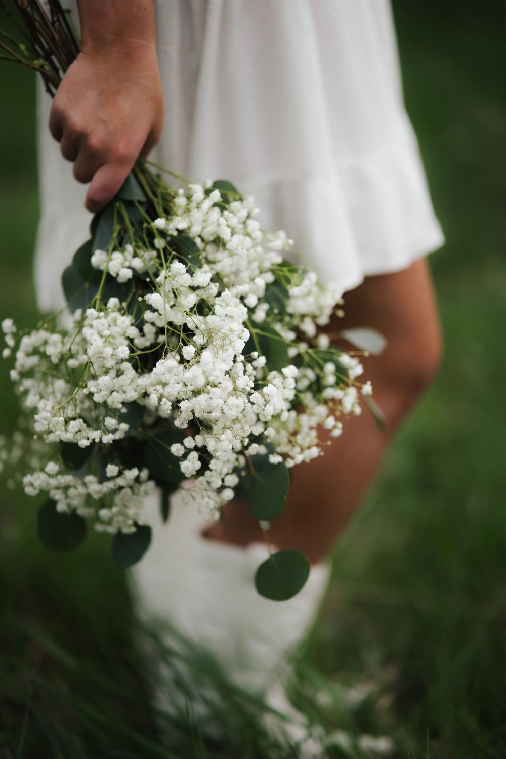 Close-up of hand holding white gypsophila and eucalyptus bouquet against white dress in green grass.
