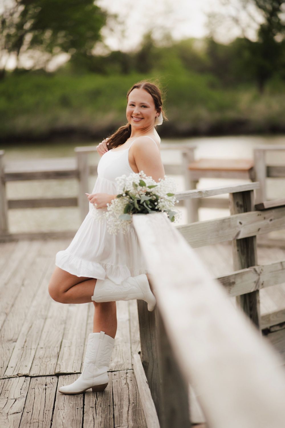 Smiling young woman in white dress and cowboy boots holding flower bouquet on wooden dock.
