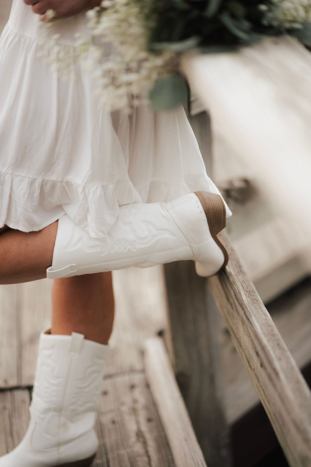 Detail shot of white cowboy boots under ruffled white dress on wooden bridge.