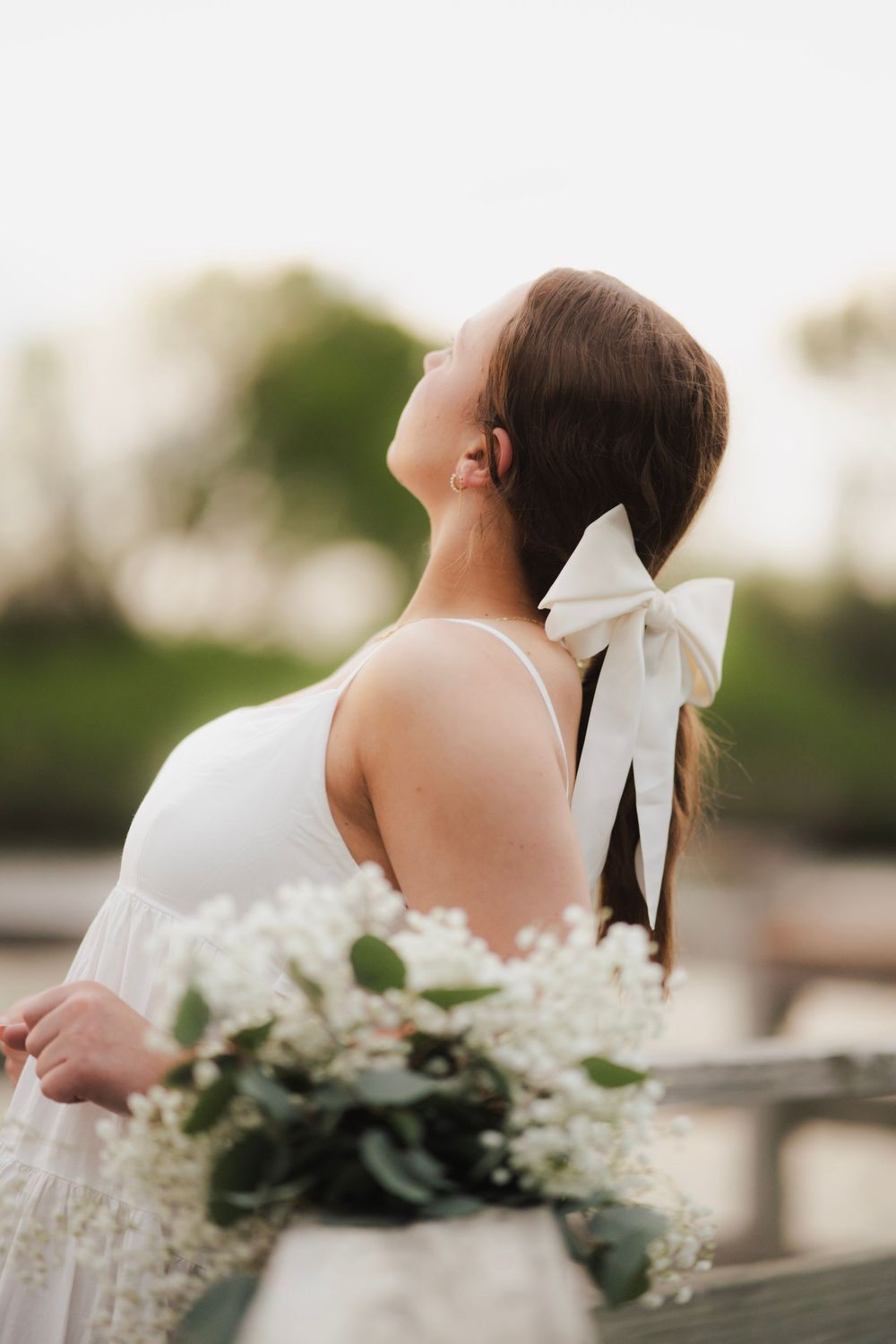 Young woman with white bow in ponytail holding floral bouquet on outdoor wooden dock.
