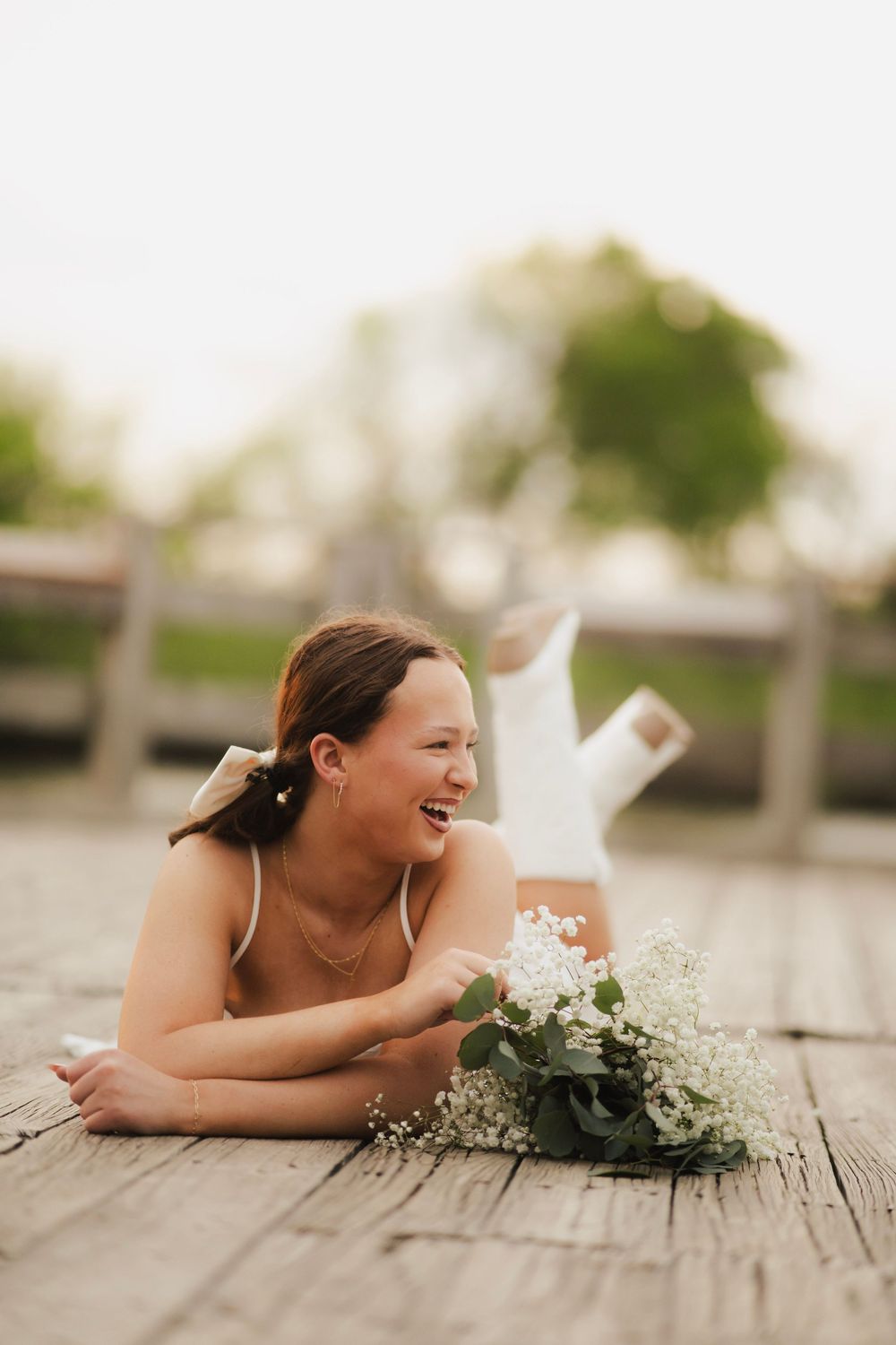 Laughing young woman lying on wooden dock holding white floral bouquet outdoors.
