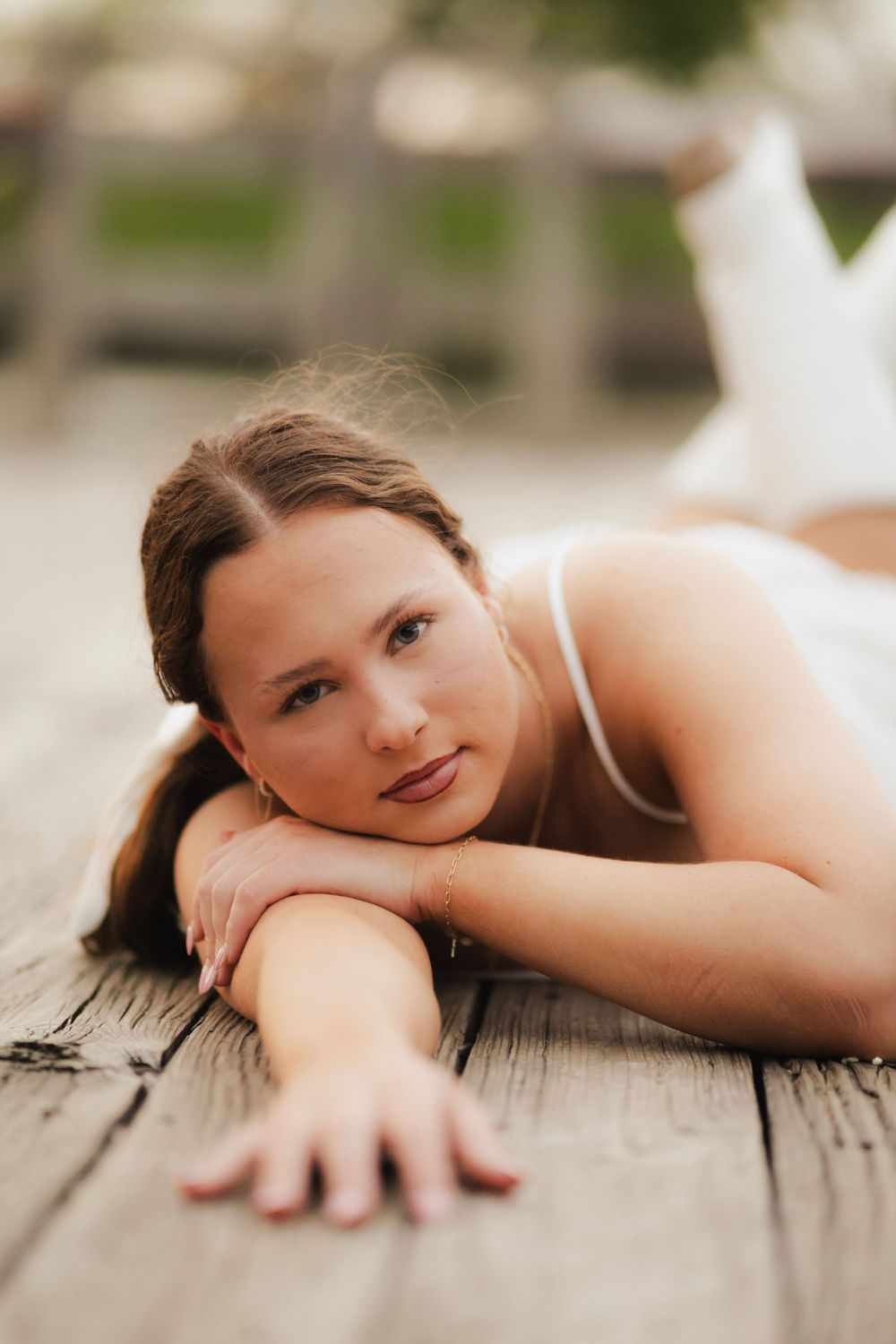 Young woman in white dress lying on weathered wooden dock, resting chin on hands, gazing at camera.