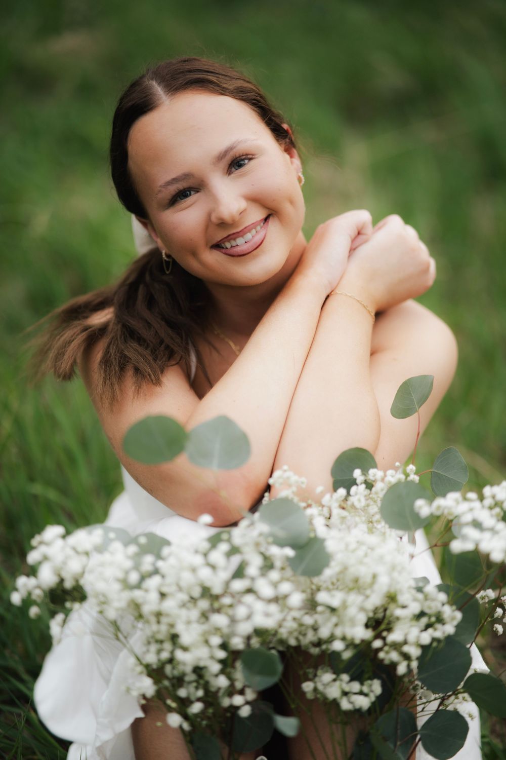 Smiling young woman in white dress holding bouquet of white gypsophila and eucalyptus while sitting in green grass.