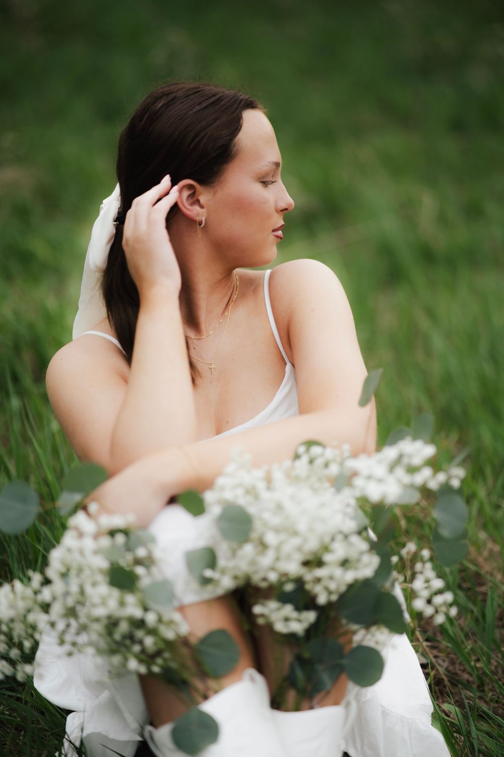 Young woman in white dress with white bow adjusting hair while sitting with floral bouquet in green grass.