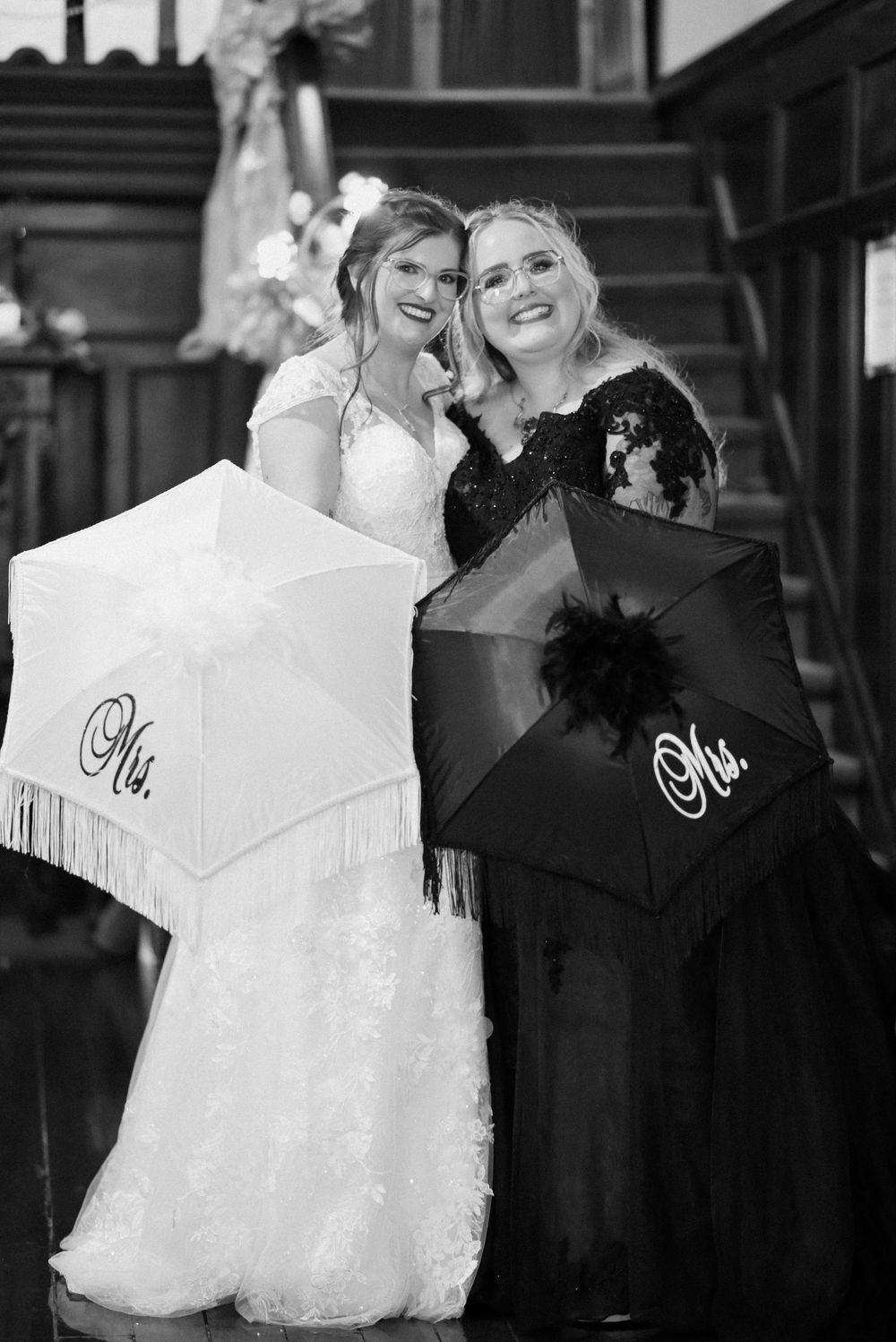 Two brides pose with Mrs. parasols in black and white wedding photo on elegant staircase.