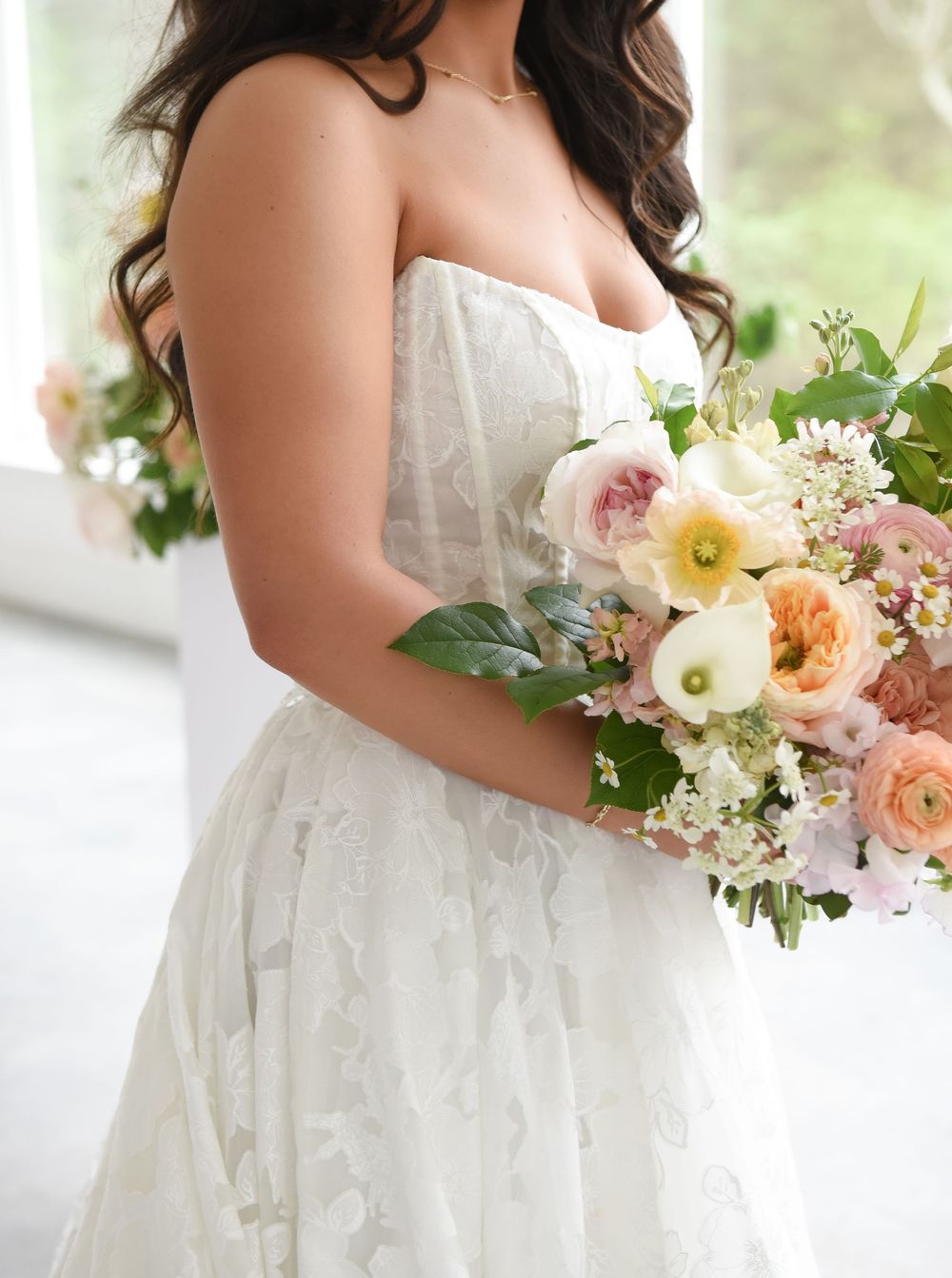 Bride in strapless white lace gown holding a lush pastel bouquet of peach, pink, and white flowers with greenery.