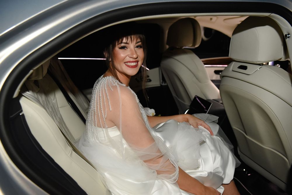 Smiling bride in white gown seated in luxury car backseat, looking over shoulder for wedding photo.