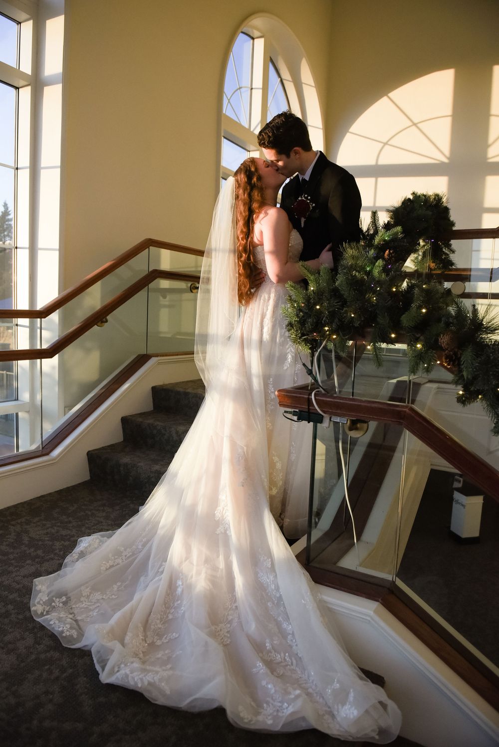 Bride and groom share a kiss on elegant staircase, bride wearing lace mermaid gown with veil, holiday greenery nearby.