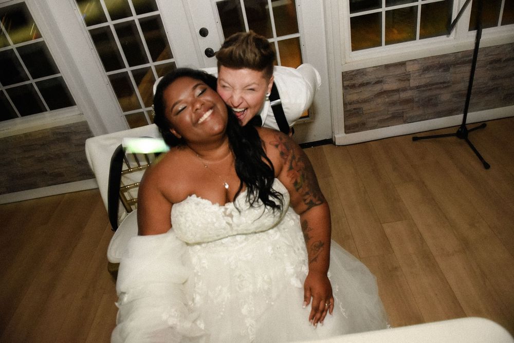 Two brides in white wedding gowns laughing joyfully together at their wedding reception venue.