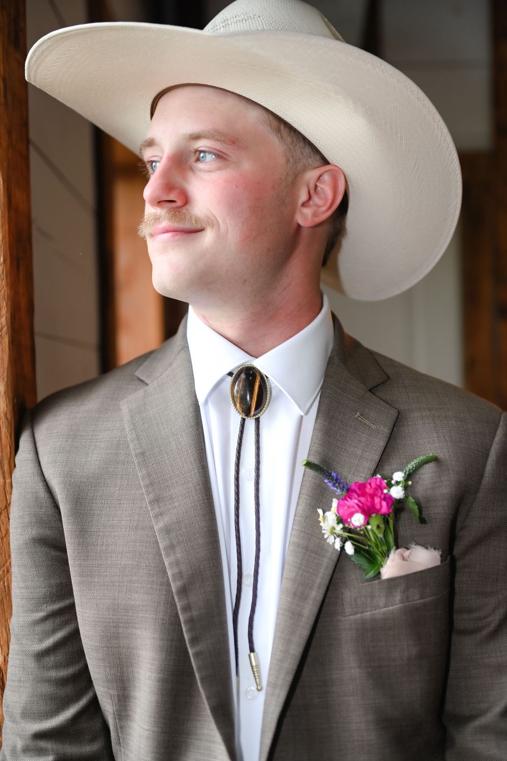 Three groomsmen in gray suits and white cowboy hats with boutonnières stand in a rustic wooden setting.