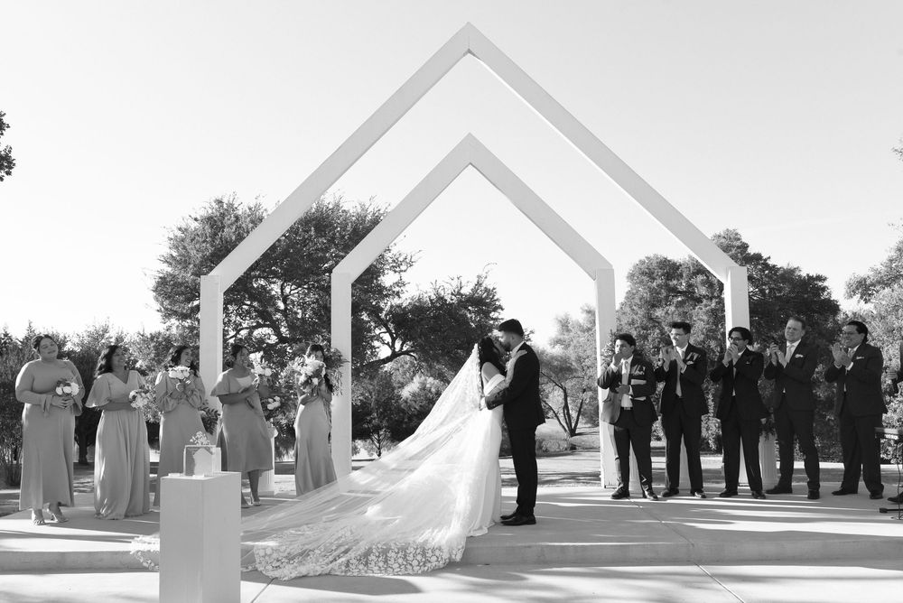 Black and white wedding ceremony photo of couple kissing under geometric arch with wedding party watching outdoors.