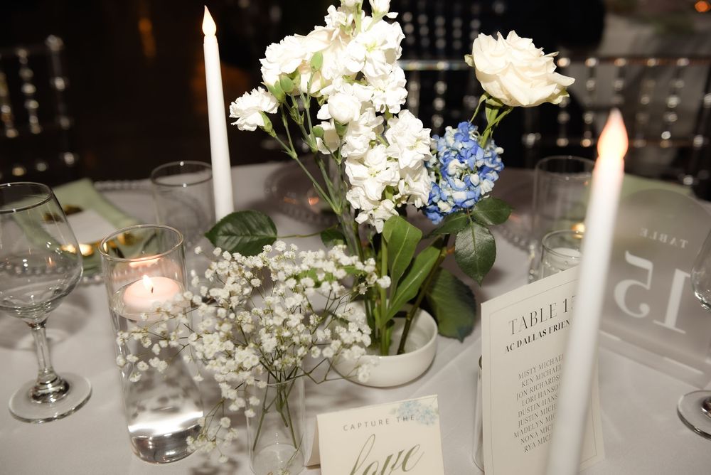 Elegant wedding table centerpiece with white roses, blue hydrangeas, and baby's breath in a white vase with candles.