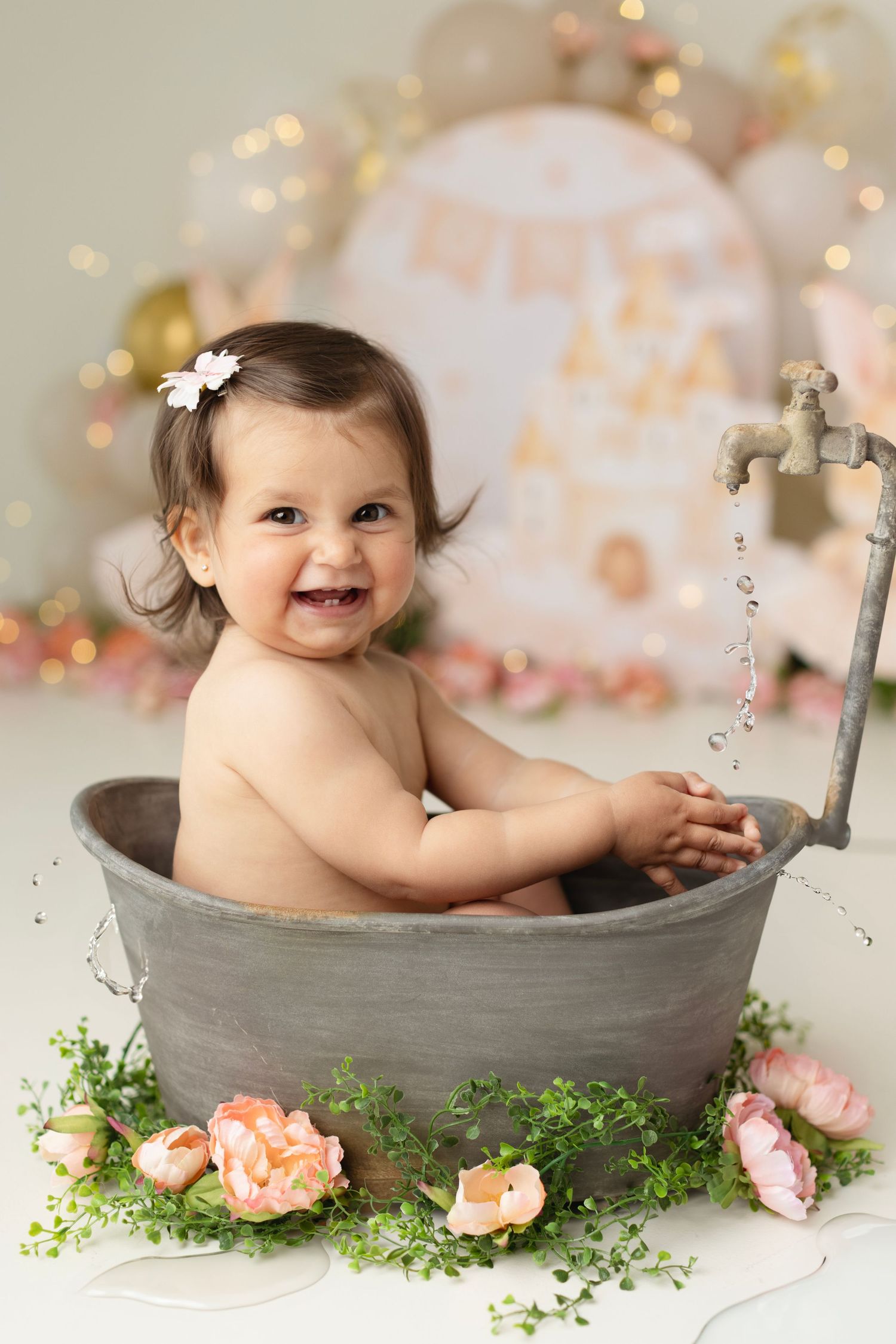 Smiling baby girl sitting in a vintage metal tub with a faucet, surrounded by pink flowers and greenery. Elizabeth Lang Photography Surrey