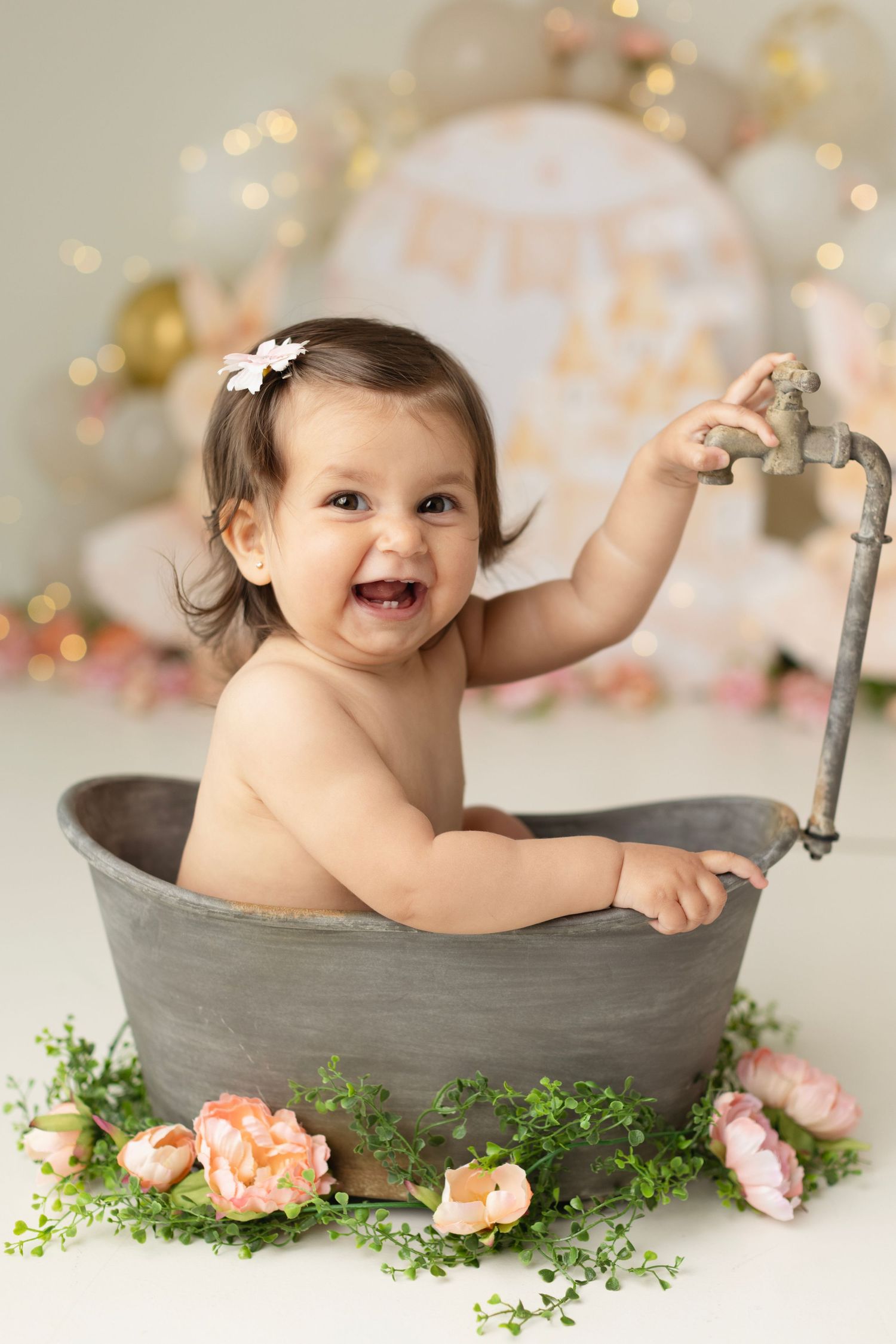Smiling baby girl sitting in a vintage metal tub surrounded by pink flowers and greenery during a first birthday photo shoot. Elizabeth Lang