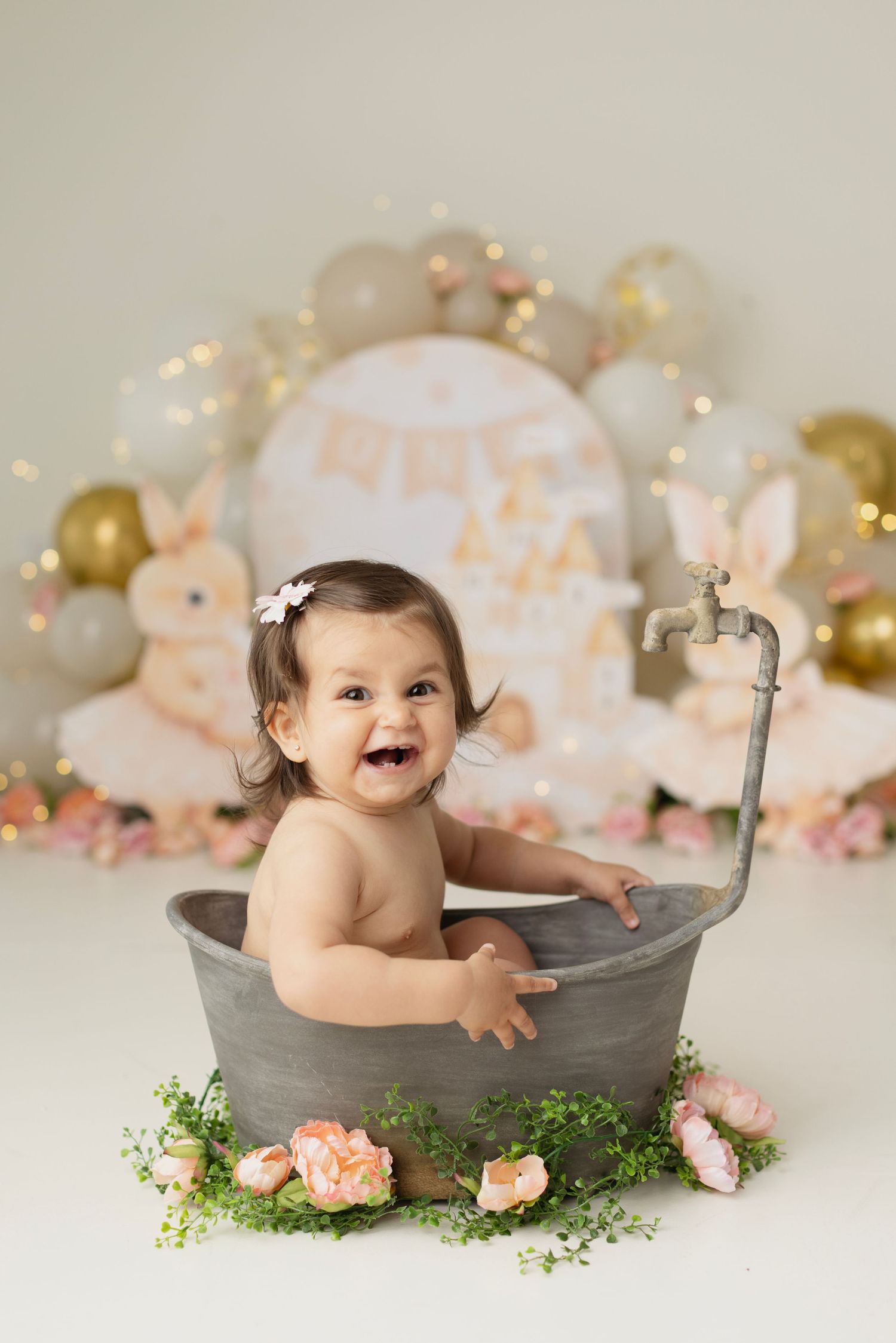 A laughing baby sits in a rustic tin tub surrounded by flowers, with a bunny-themed birthday backdrop behind her. Elizabeth Lang Photography