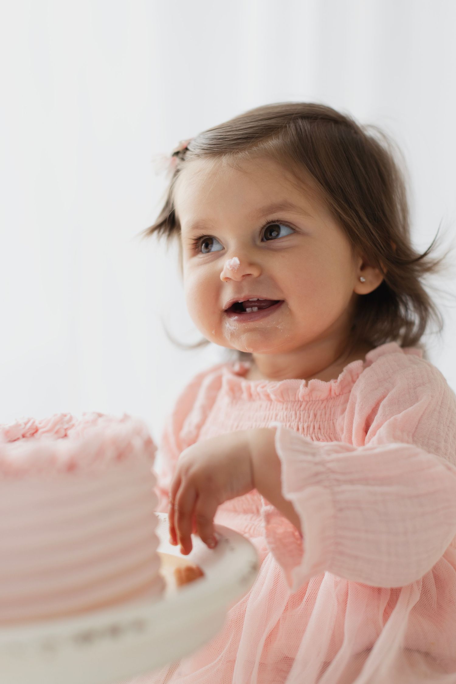 Smiling baby girl in pink dress touching a pink birthday cake during her first birthday cake smash photo session. Elizabeth Lang Photography