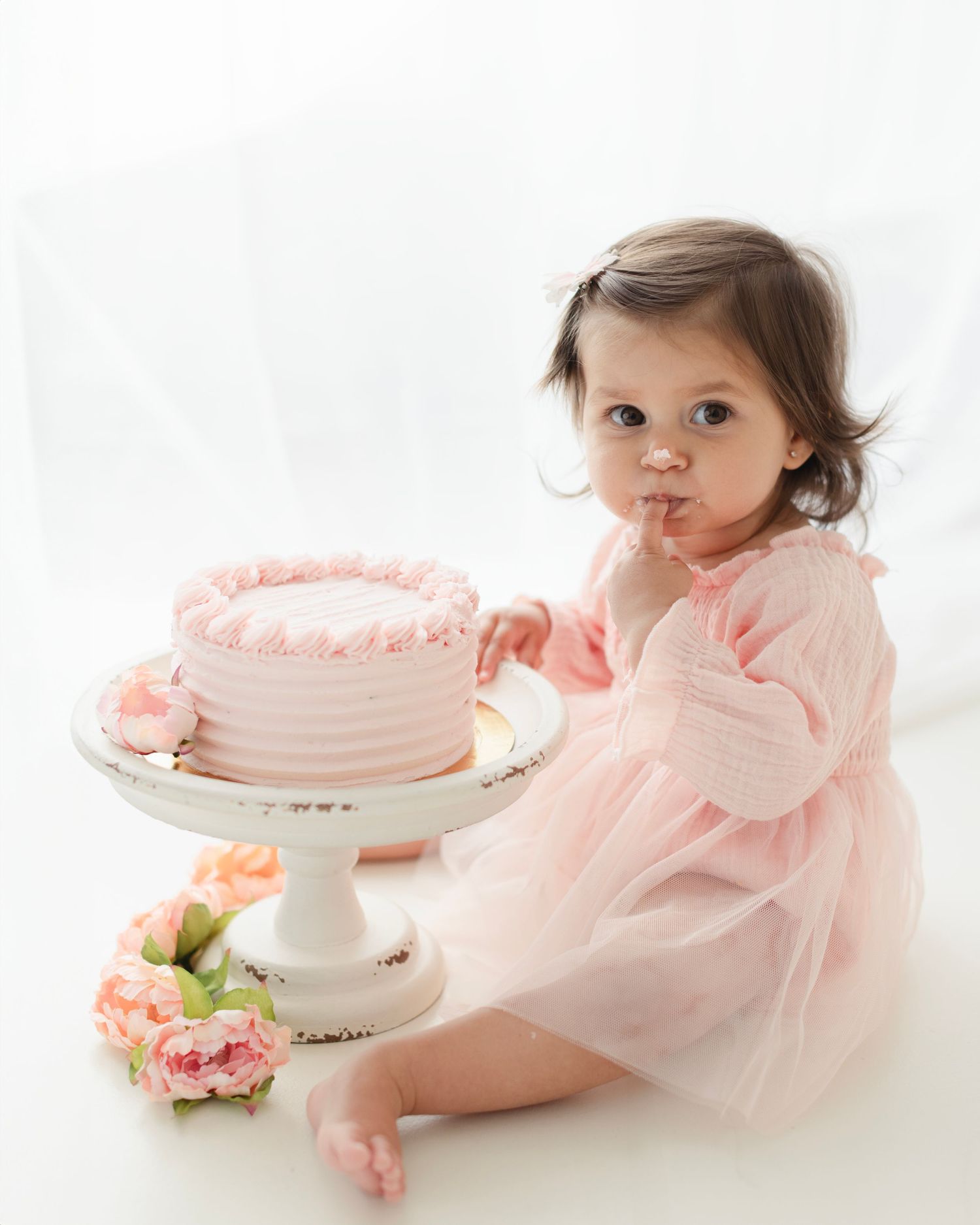Baby girl in pink dress sits beside a pink birthday cake on a white cake stand during a first birthday cake smash photo session. Elizabeth