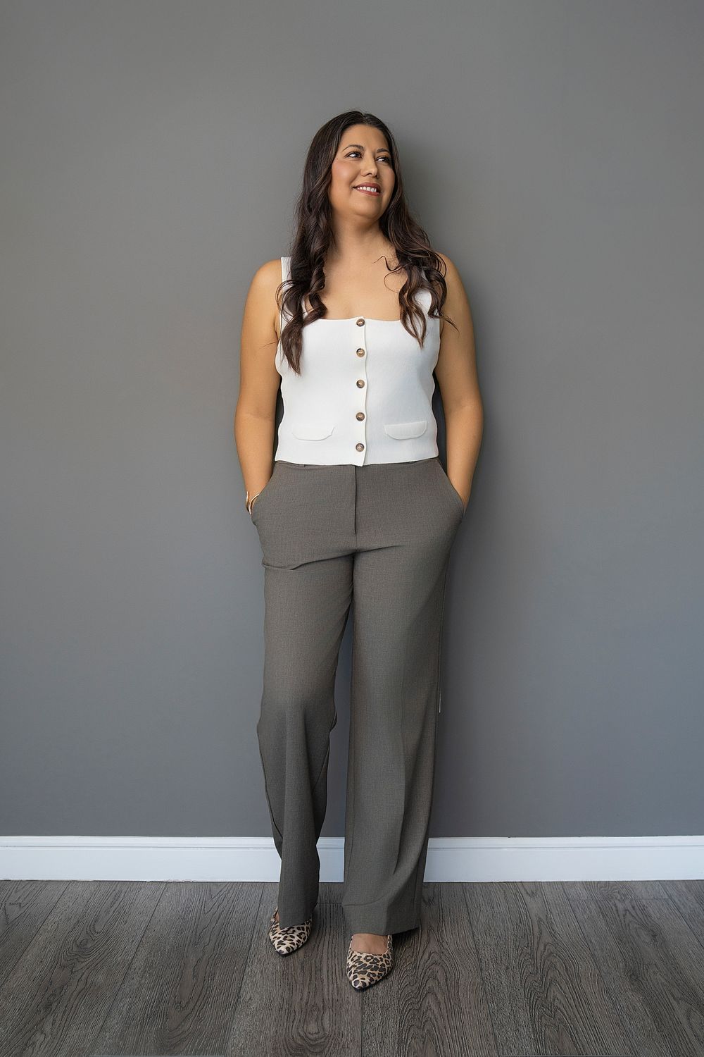 Woman in white button top and olive wide-leg trousers with leopard flats, standing against gray wall.