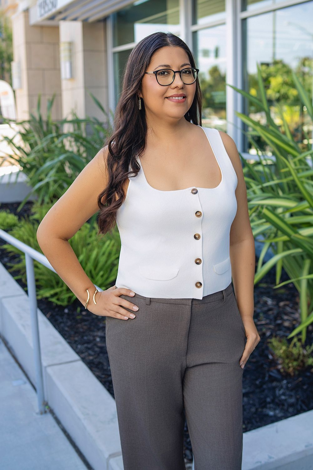 Professional woman with glasses in white top and gray pants standing outside modern building with greenery.