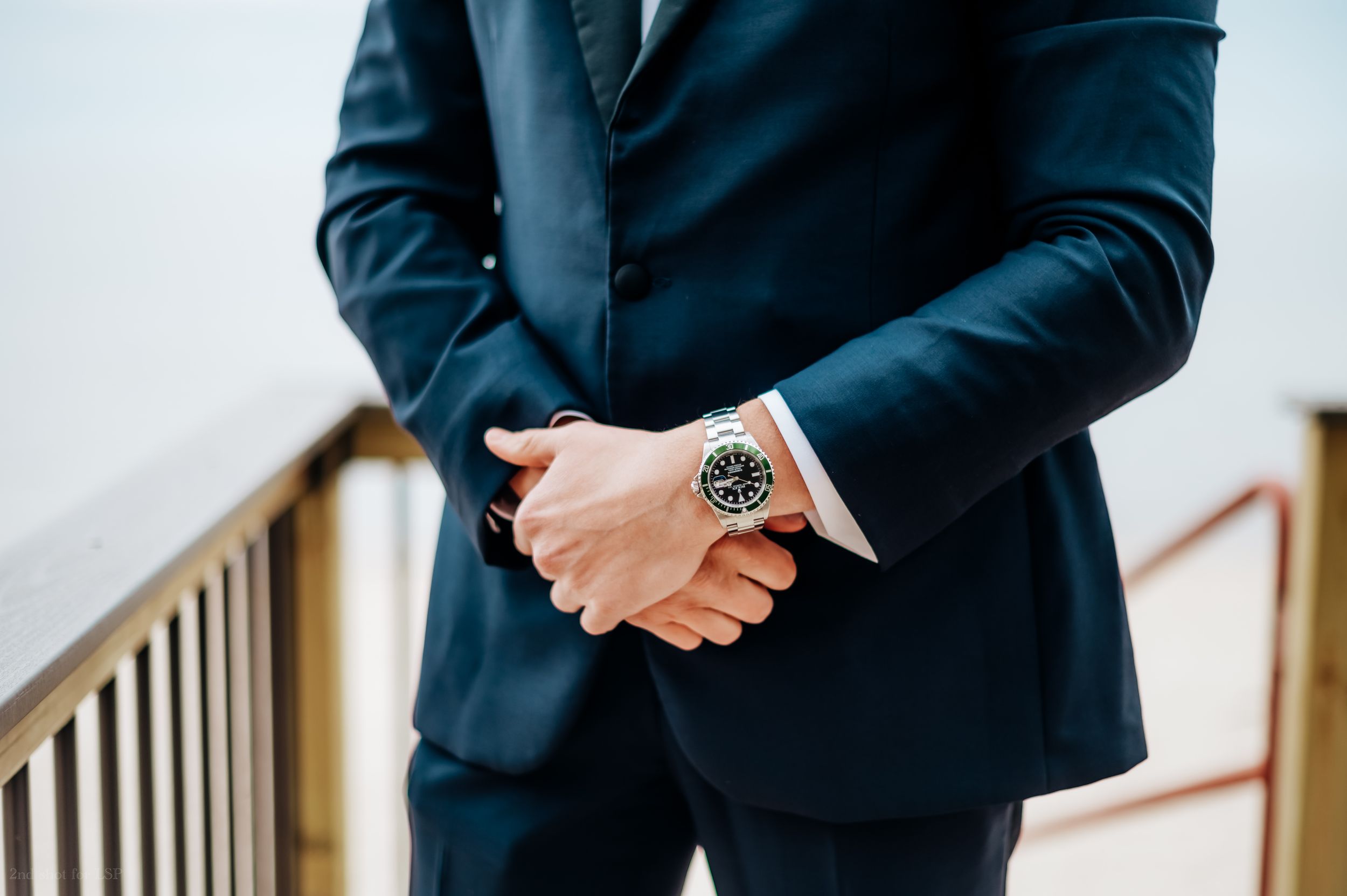 Man in navy blue suit with watch clasping hands, formal business attire close-up shot.