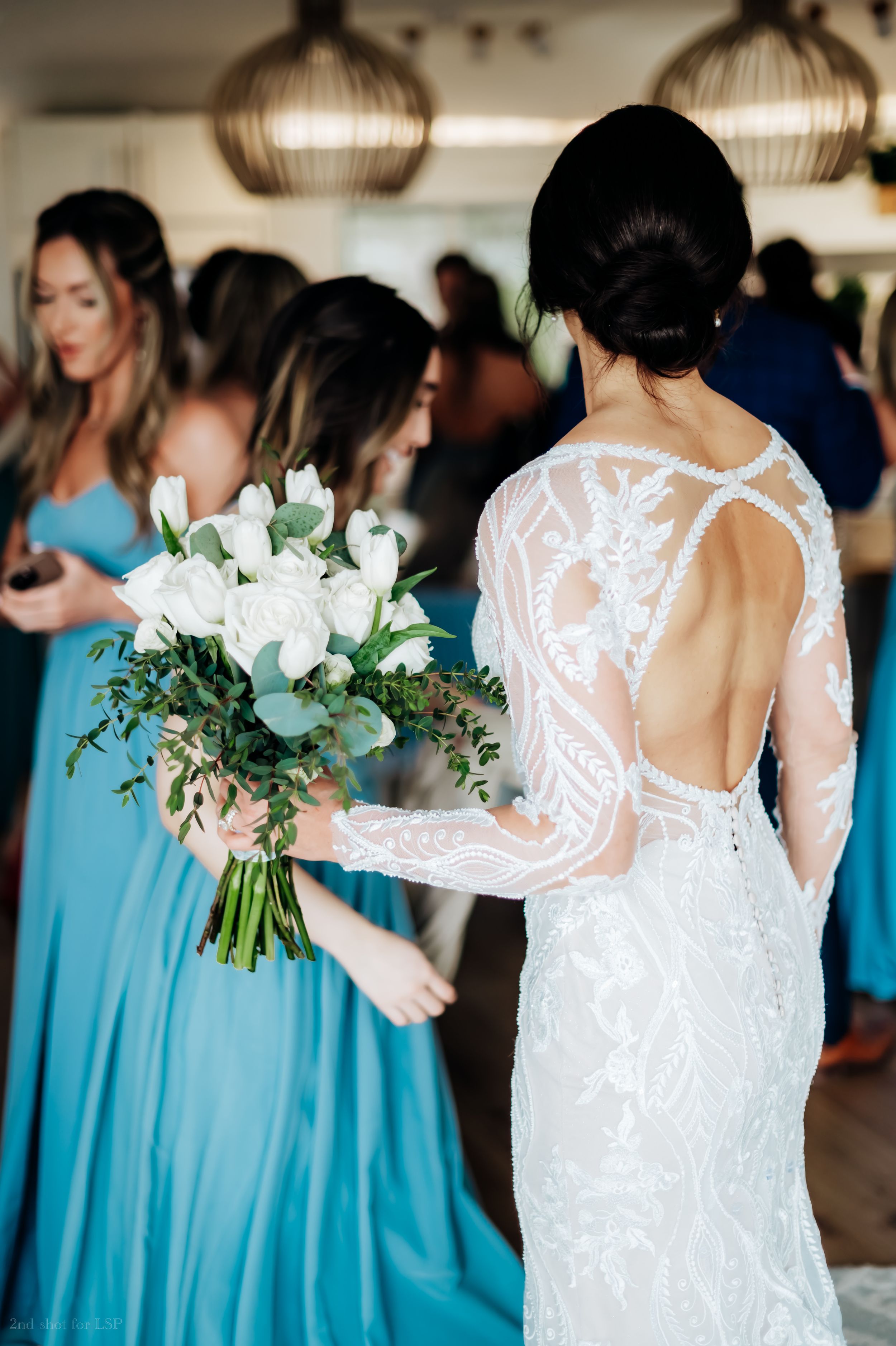 Bride in lace backless gown holds white bouquet with bridesmaids in blue dresses behind her.