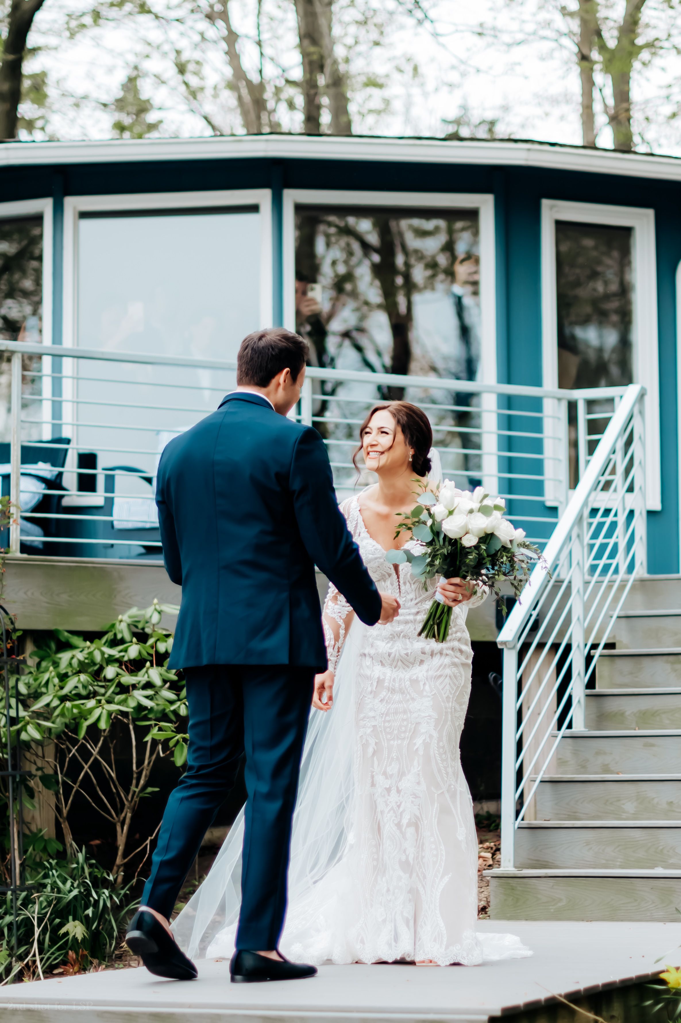 Bride in lace gown and groom in navy suit hold hands on steps outside a teal modern building.