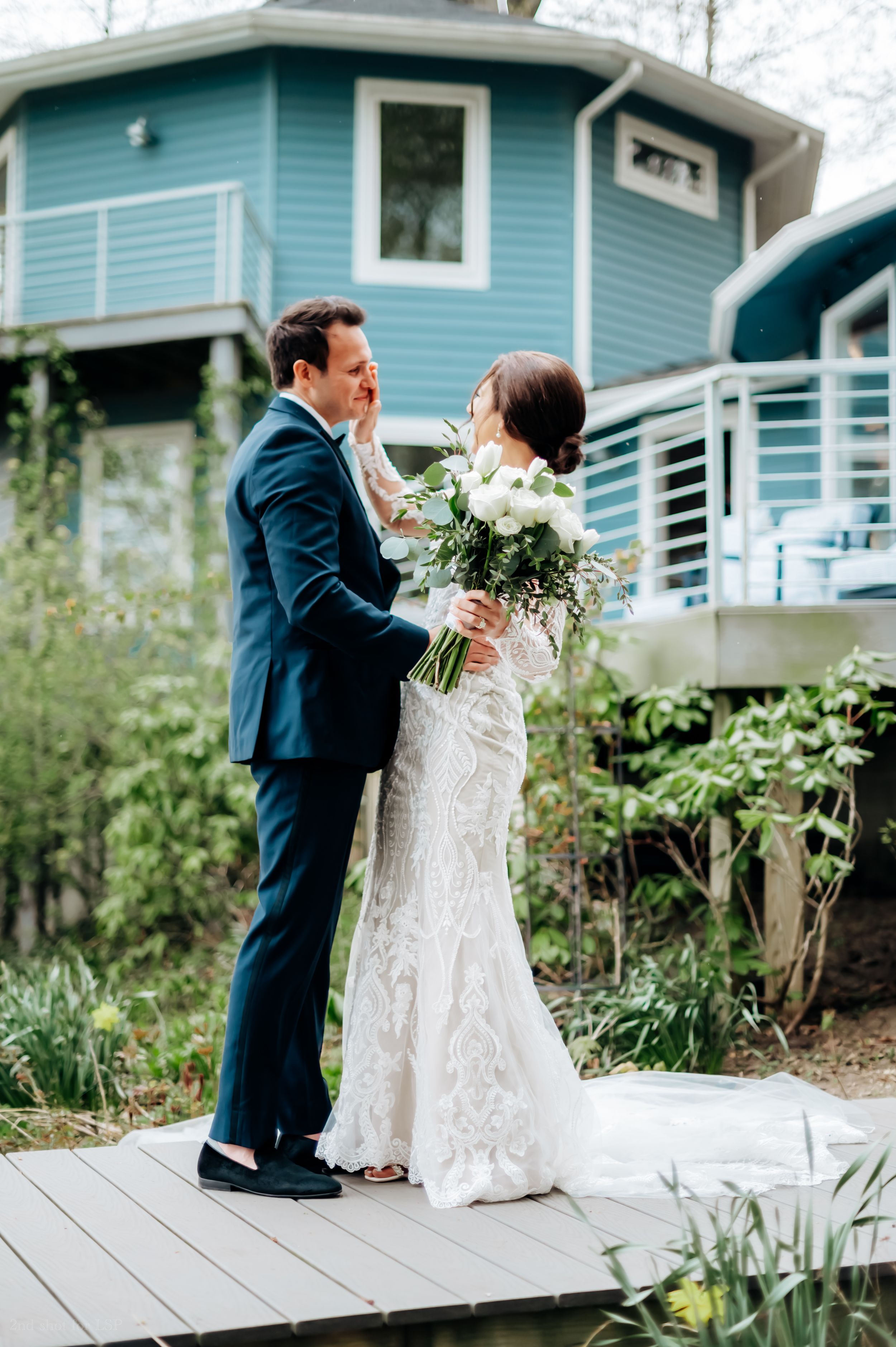 Bride and groom share a tender first look moment outdoors, with bride holding white bouquet near a blue house.