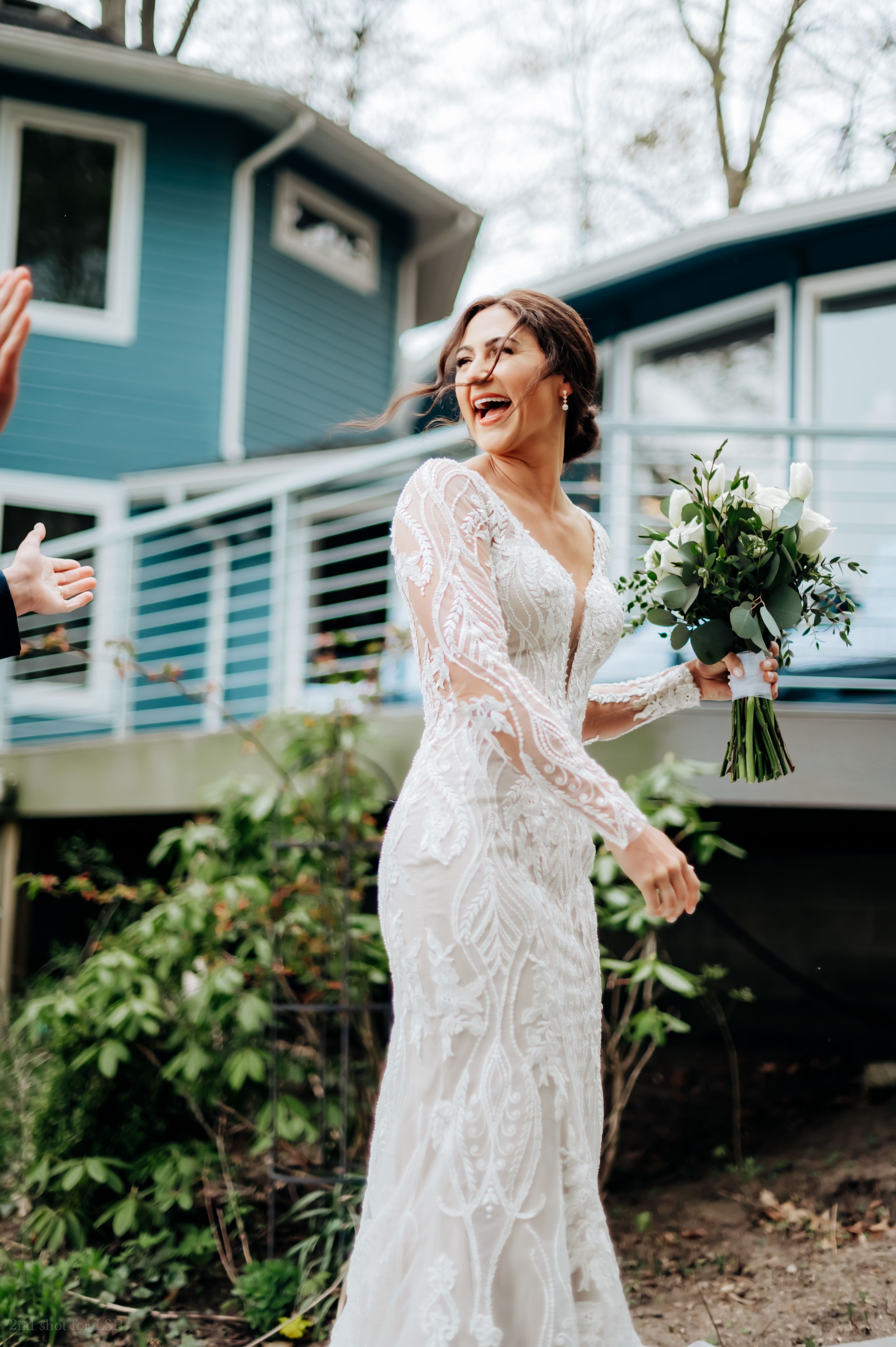 Joyful bride in long-sleeve lace wedding gown laughs while holding white floral bouquet outdoors.