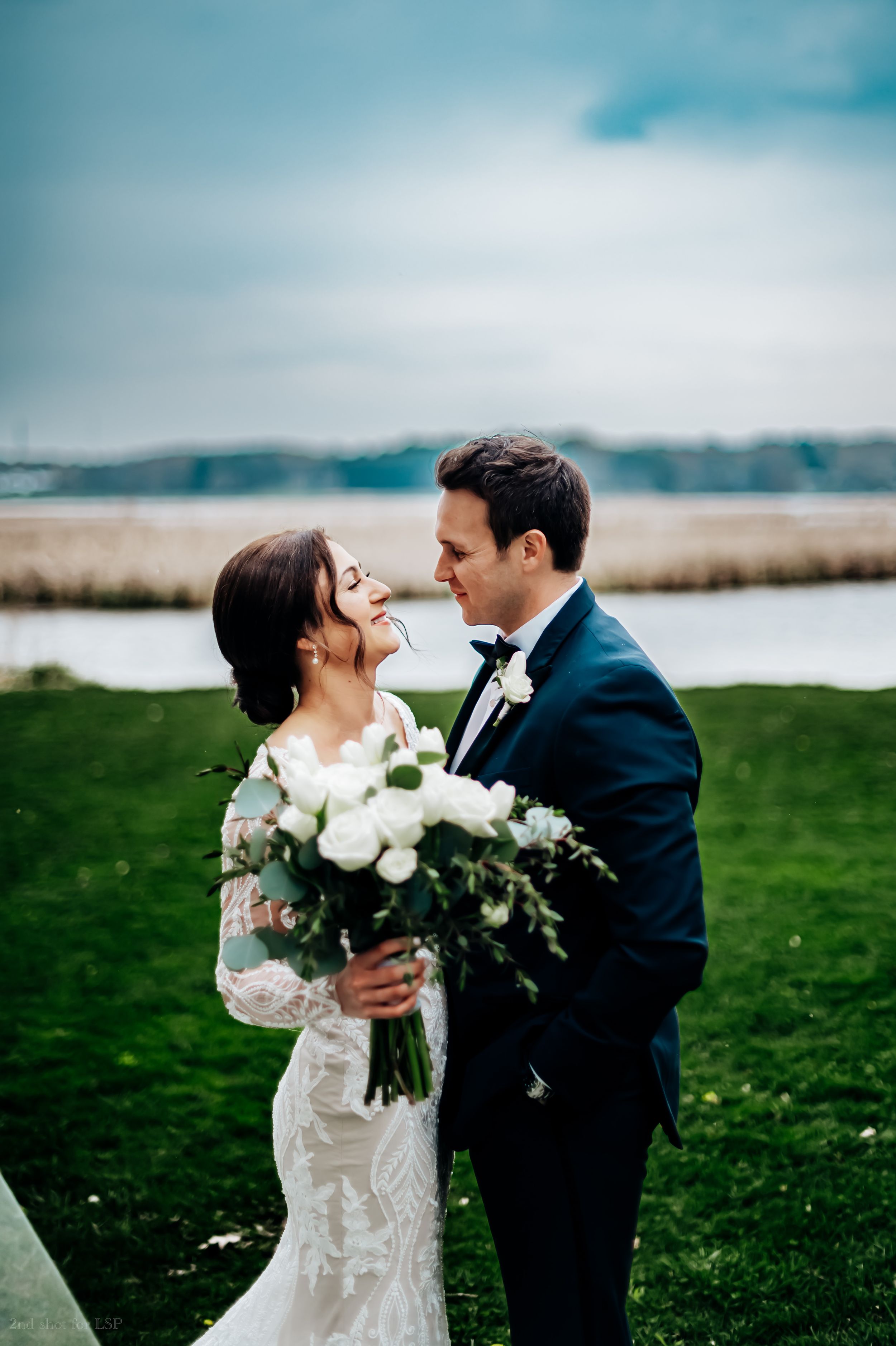 Bride and groom share romantic moment outdoors holding white rose bouquet by waterfront.