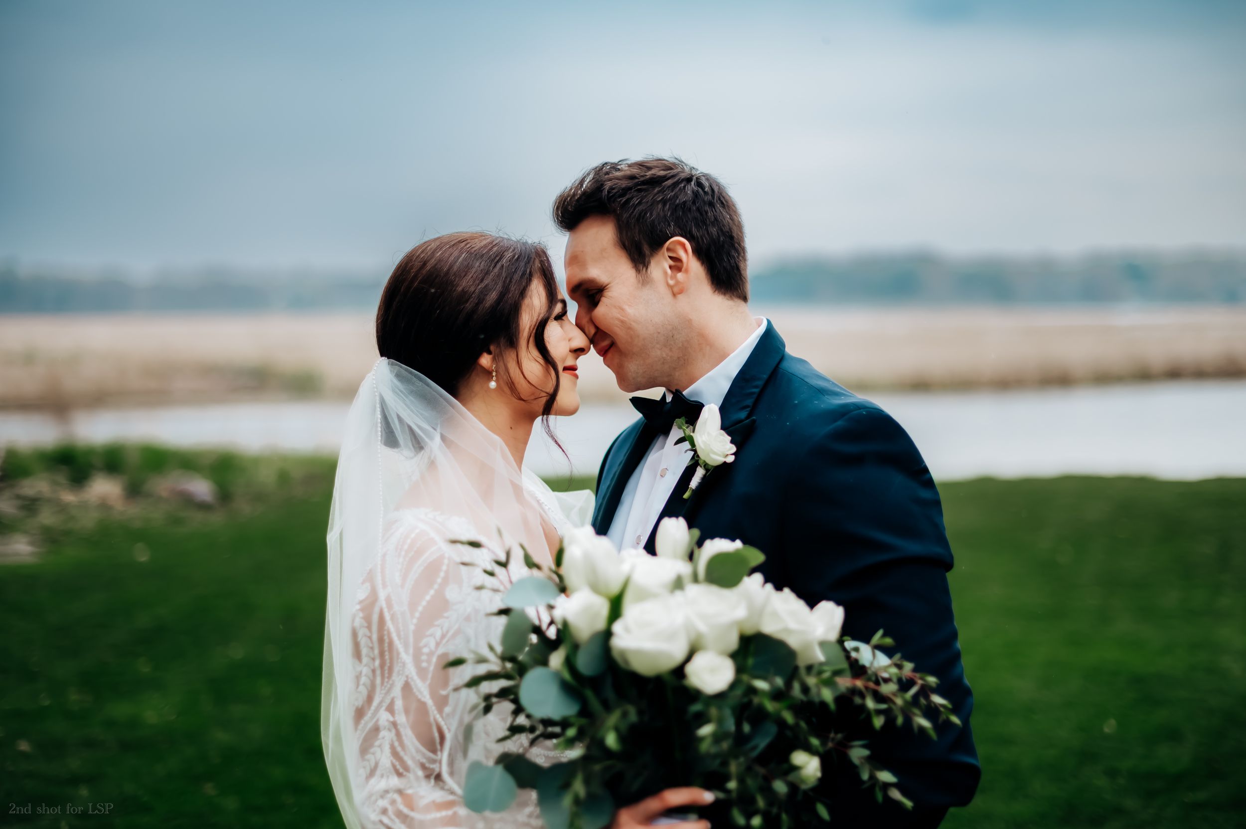 Bride and groom share romantic moment outdoors, bride holding white rose bouquet with greenery, wearing veil and lace gown.