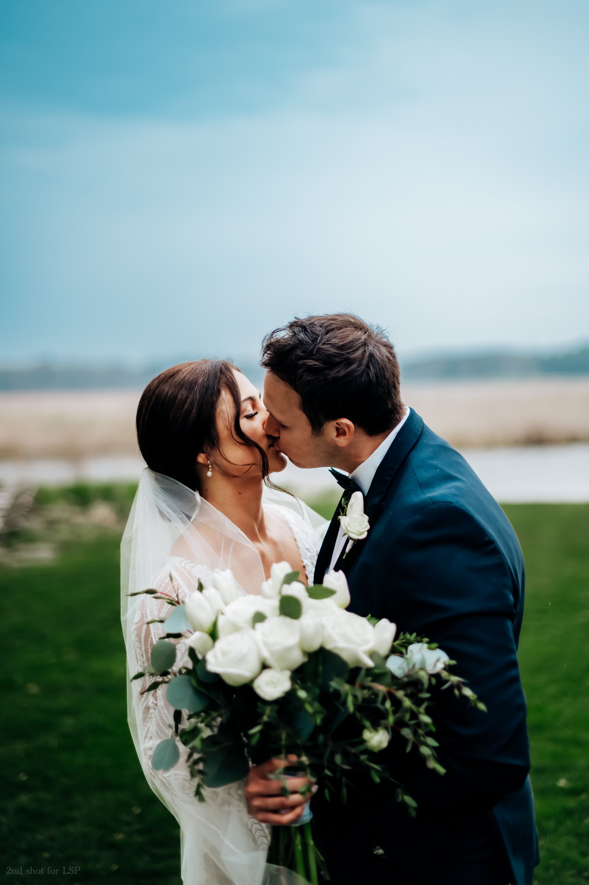 Bride and groom sharing a romantic kiss outdoors holding a white rose bouquet with greenery on their wedding day.
