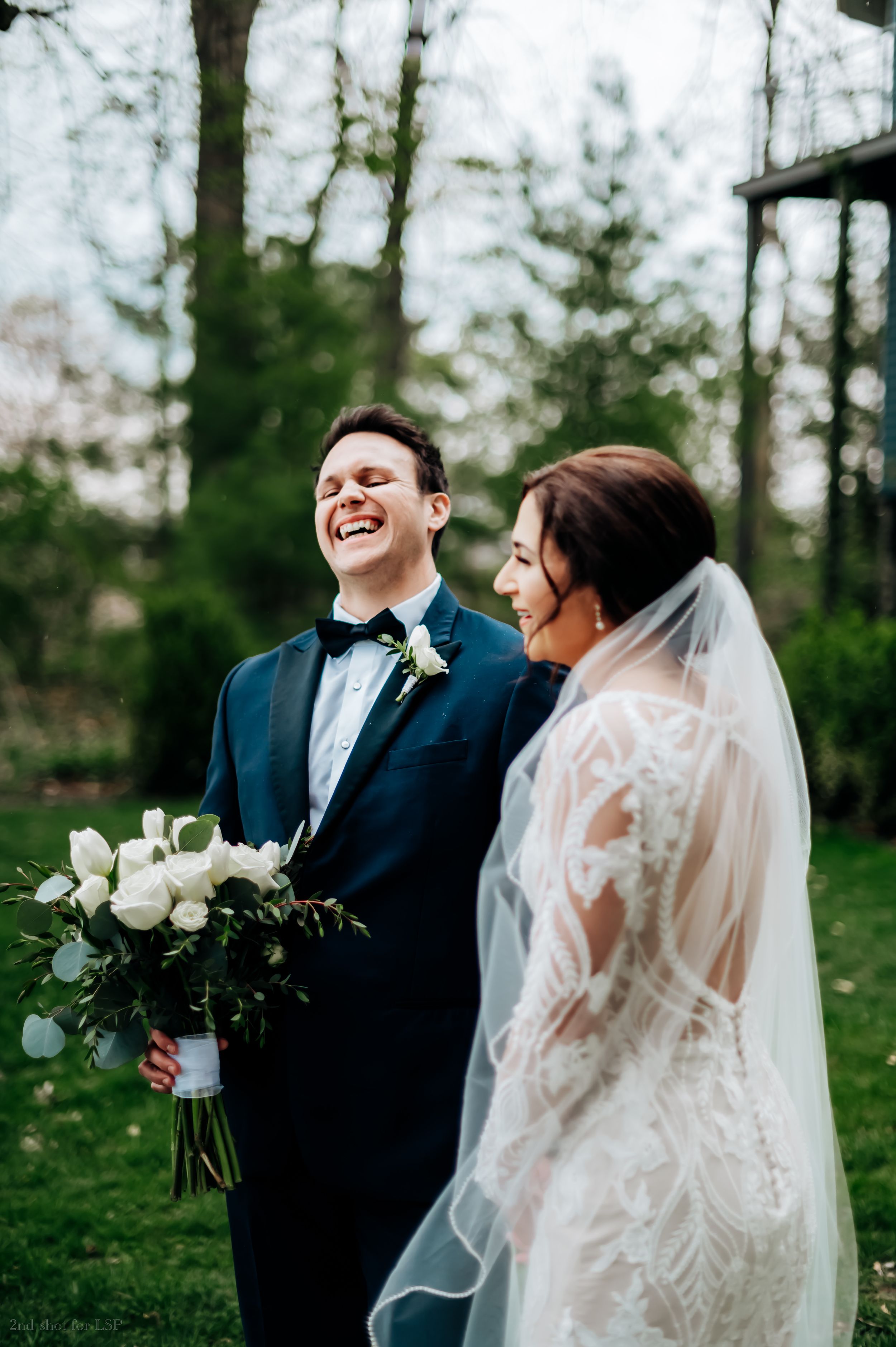 Groom in tuxedo laughing joyfully during first look with bride in lace wedding gown.