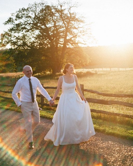 Bride and groom walk hand-in-hand on a forest path, she in a white gown with sunflowers, he in a blue suit.