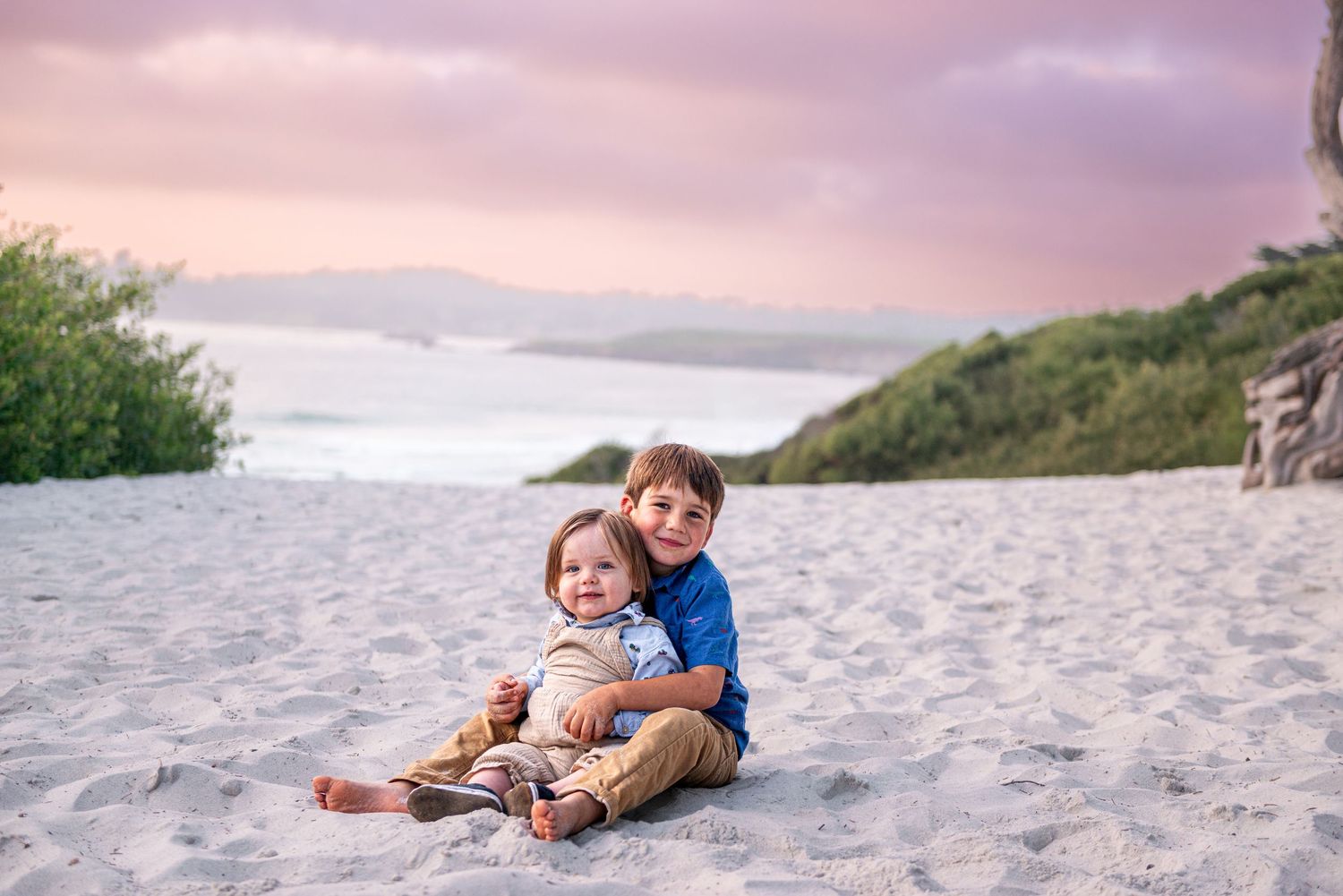 Two young brothers sit together on sandy beach at sunset, older child hugging younger sibling with ocean waves behind them.
