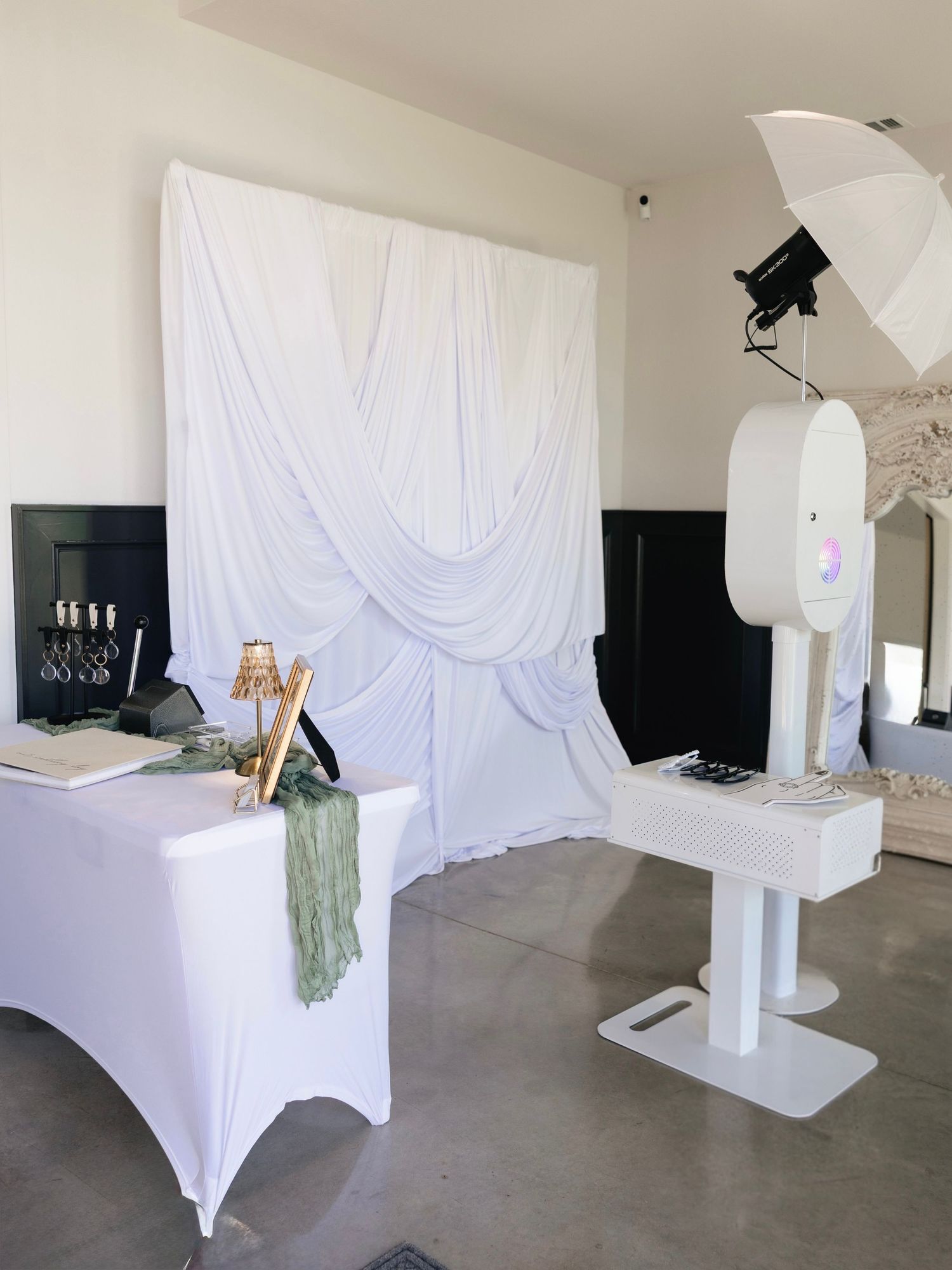 Elegant white photo booth setup with draped fabric backdrop, studio lighting, and decorated table for events.