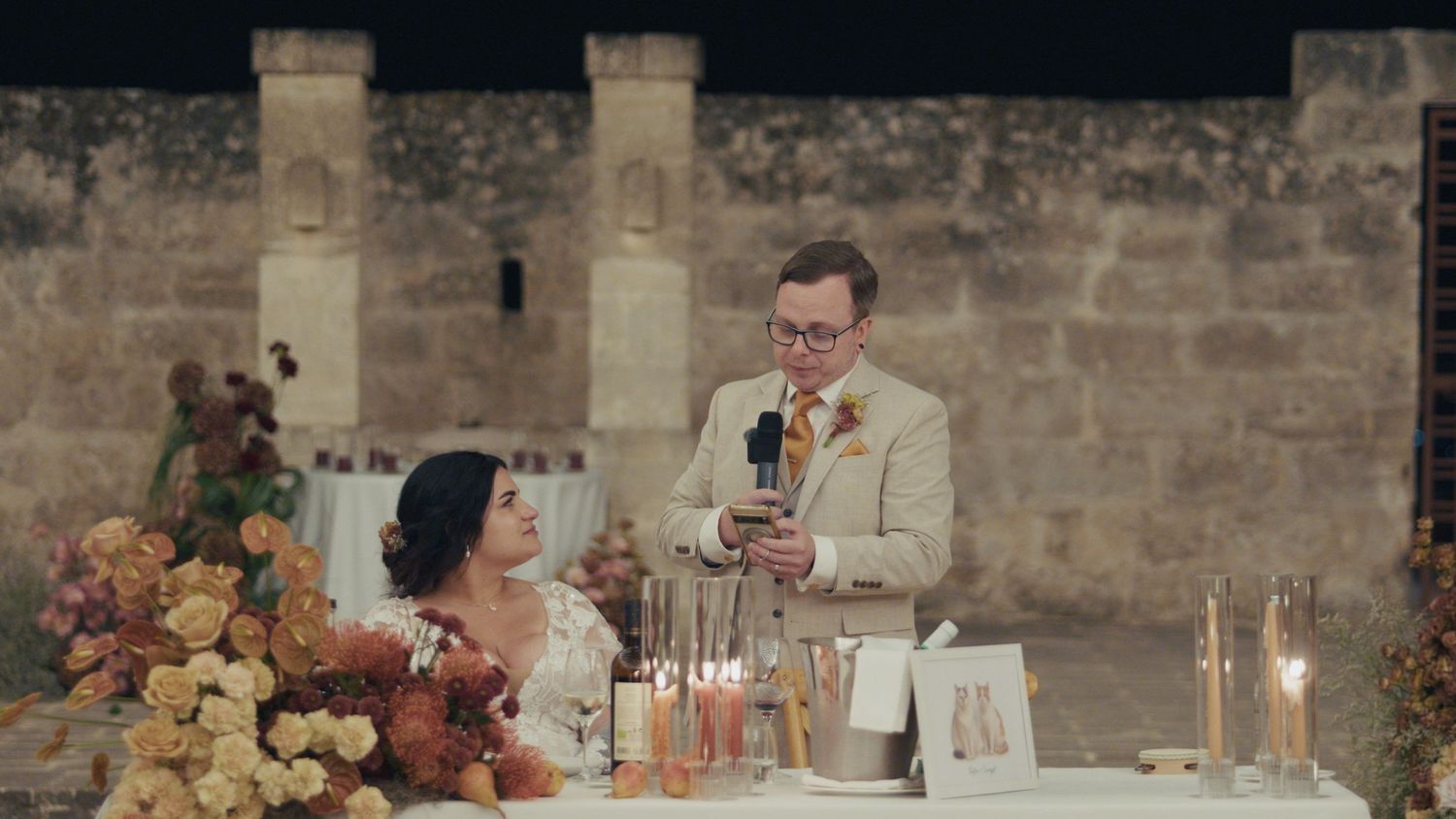 Groom reads wedding speech at candlelit reception table beside bride, rustic stone venue backdrop.