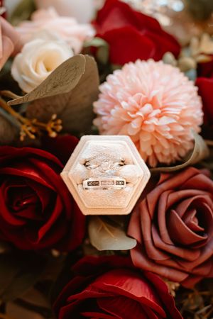 Wedding rings in hexagonal velvet box surrounded by red, pink, and mauve roses and dahlia flowers.