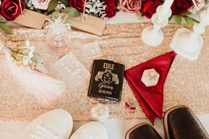Wedding flat lay with floral bouquet, personalized vow book, rings, shoes, and rose gold sequin table runner.