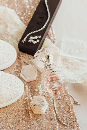 Bridal flat lay with diamond rings, pink perfume, pearl jewelry in Kay box, and lace on gold sequin fabric.