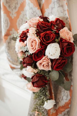 Elegant bridal bouquet featuring red, blush pink, and white roses with eucalyptus greenery against a decorative curtain.