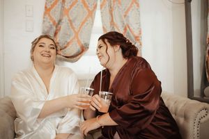 Two bridesmaids in robes toasting with drinks while laughing together during wedding preparations.