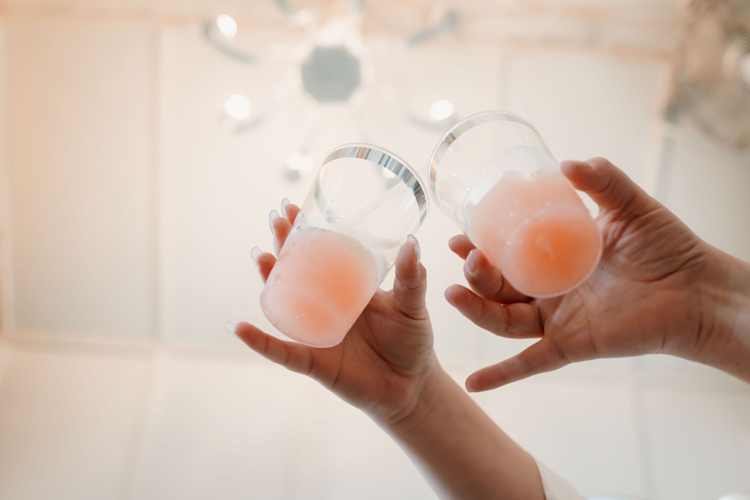 Two hands holding clear glass cups with pink bath fizz balls in a bright bathroom setting.